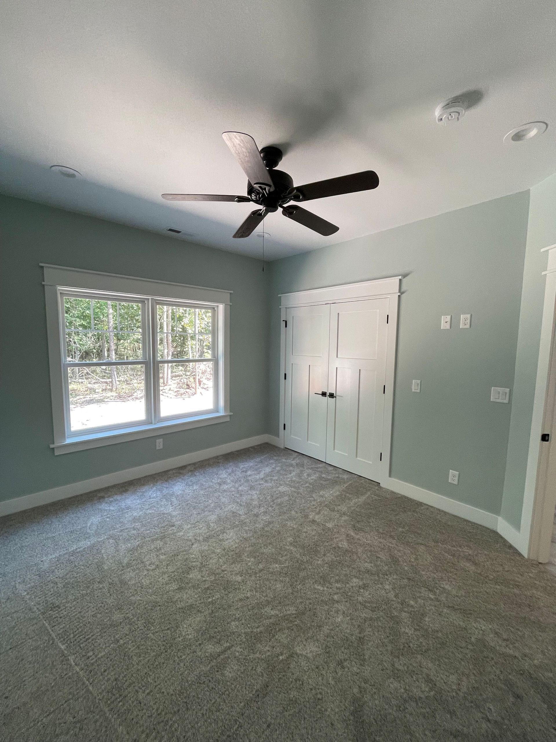 Empty bedroom with pale green walls, gray carpet, window, closet, and ceiling fan.