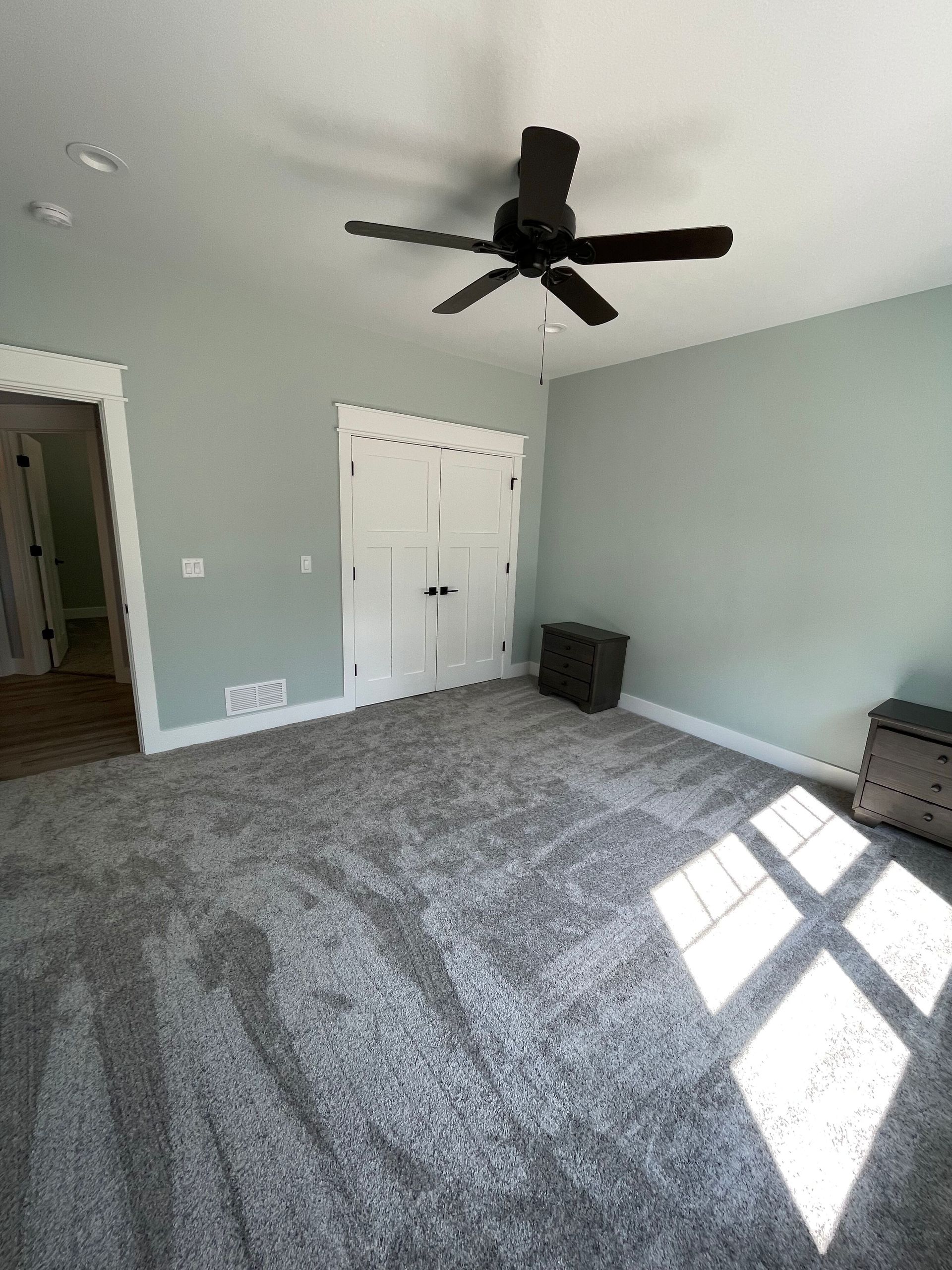 Empty gray-carpeted bedroom with light walls, ceiling fan, closet doors, and sunlight on the floor