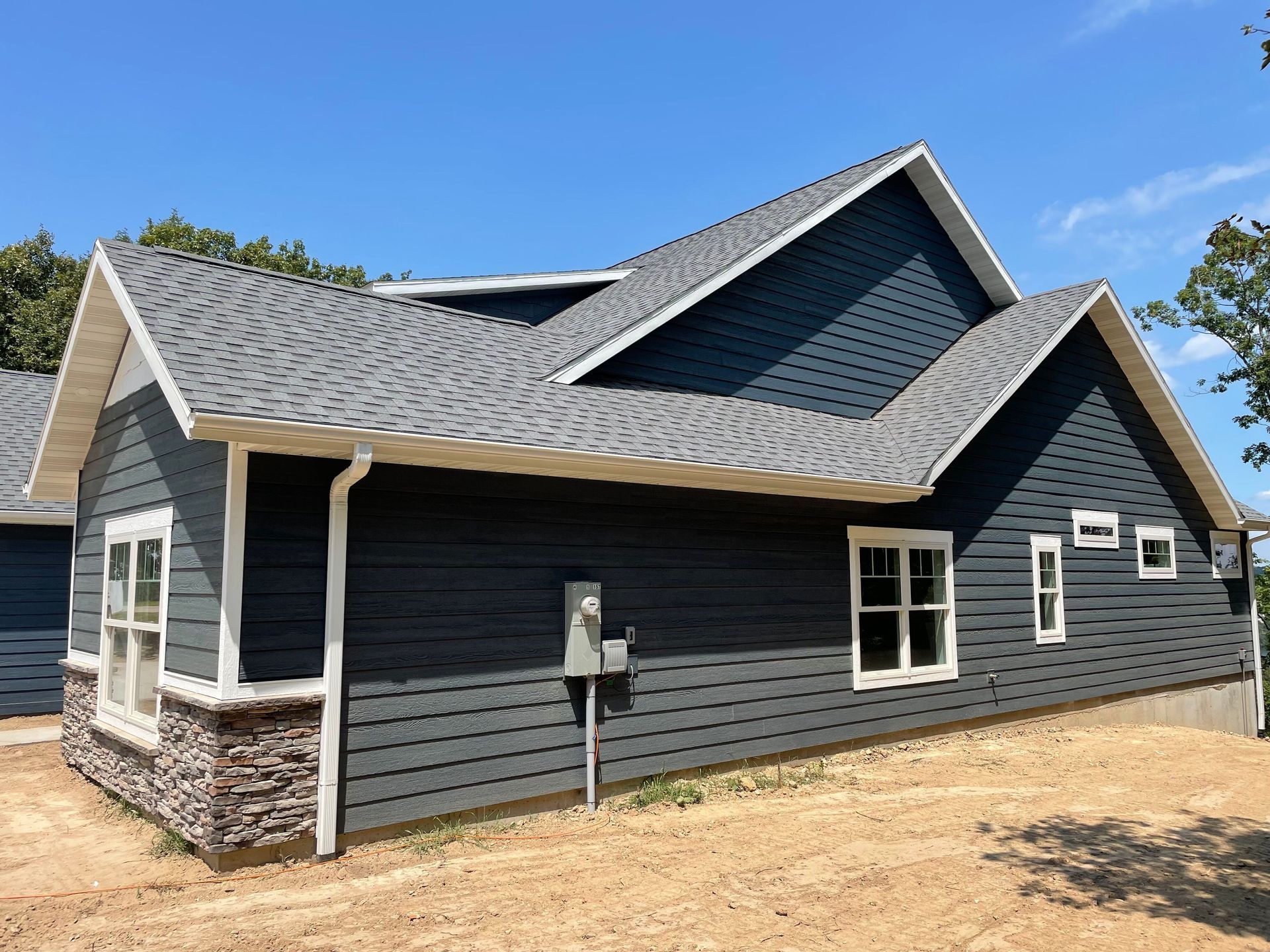 Gray house exterior with dark roof and white trim on a bare lot under a blue sky