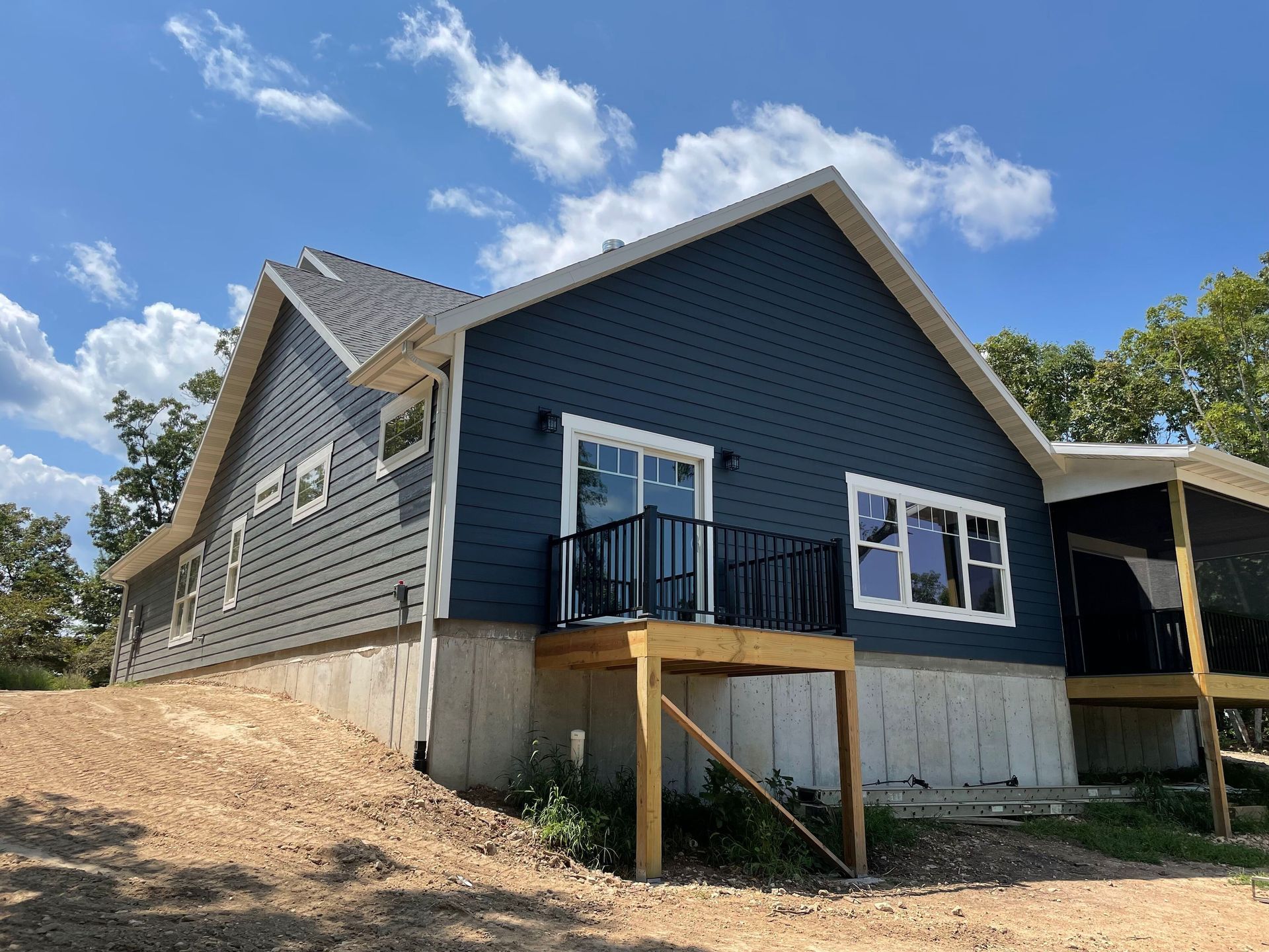 Blue house exterior with unfinished porch and concrete foundation on a dirt slope under a partly cloudy sky