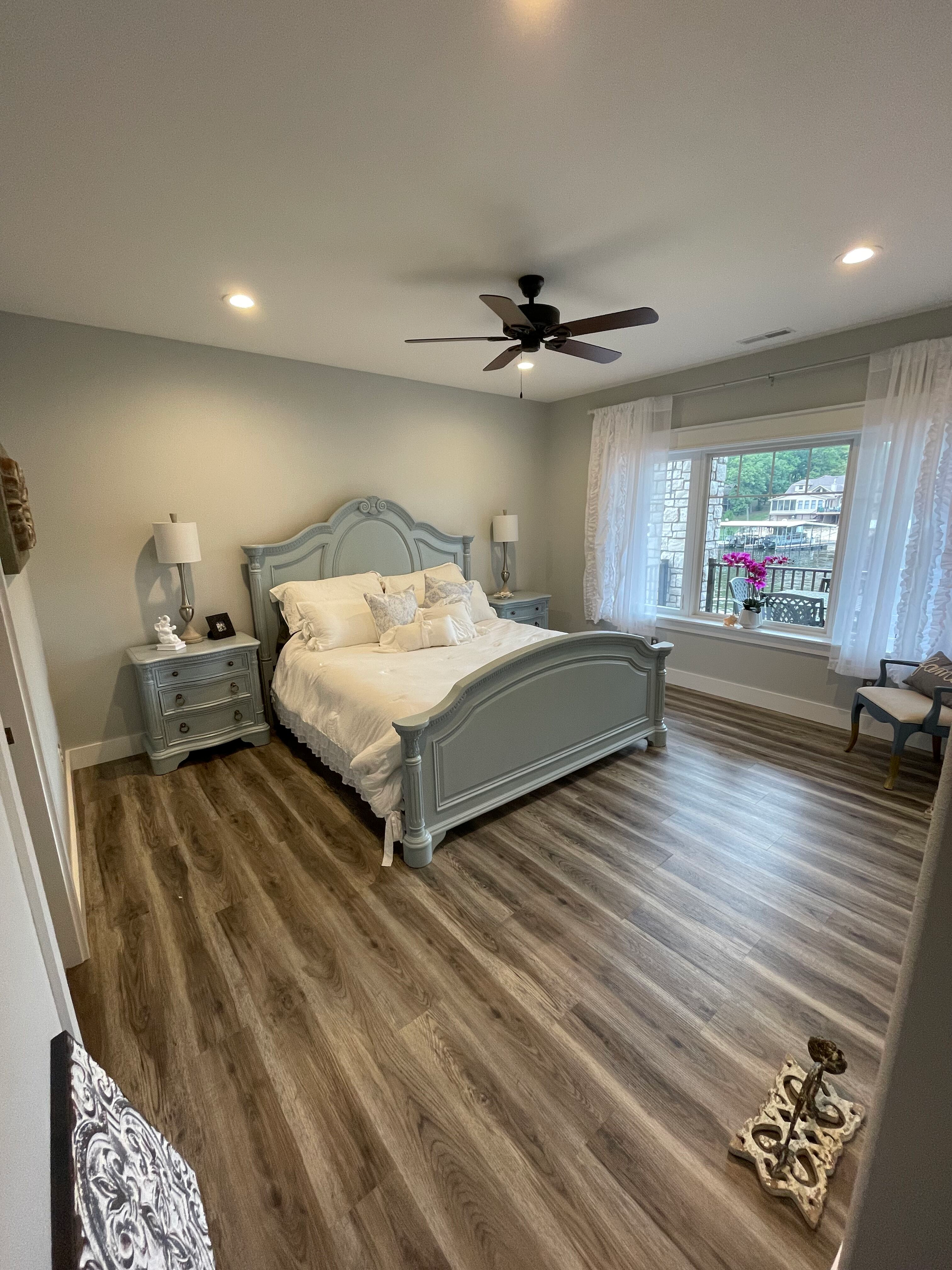 Bright bedroom with a pale blue bed, hardwood floors, ceiling fan, nightstands, and large window.