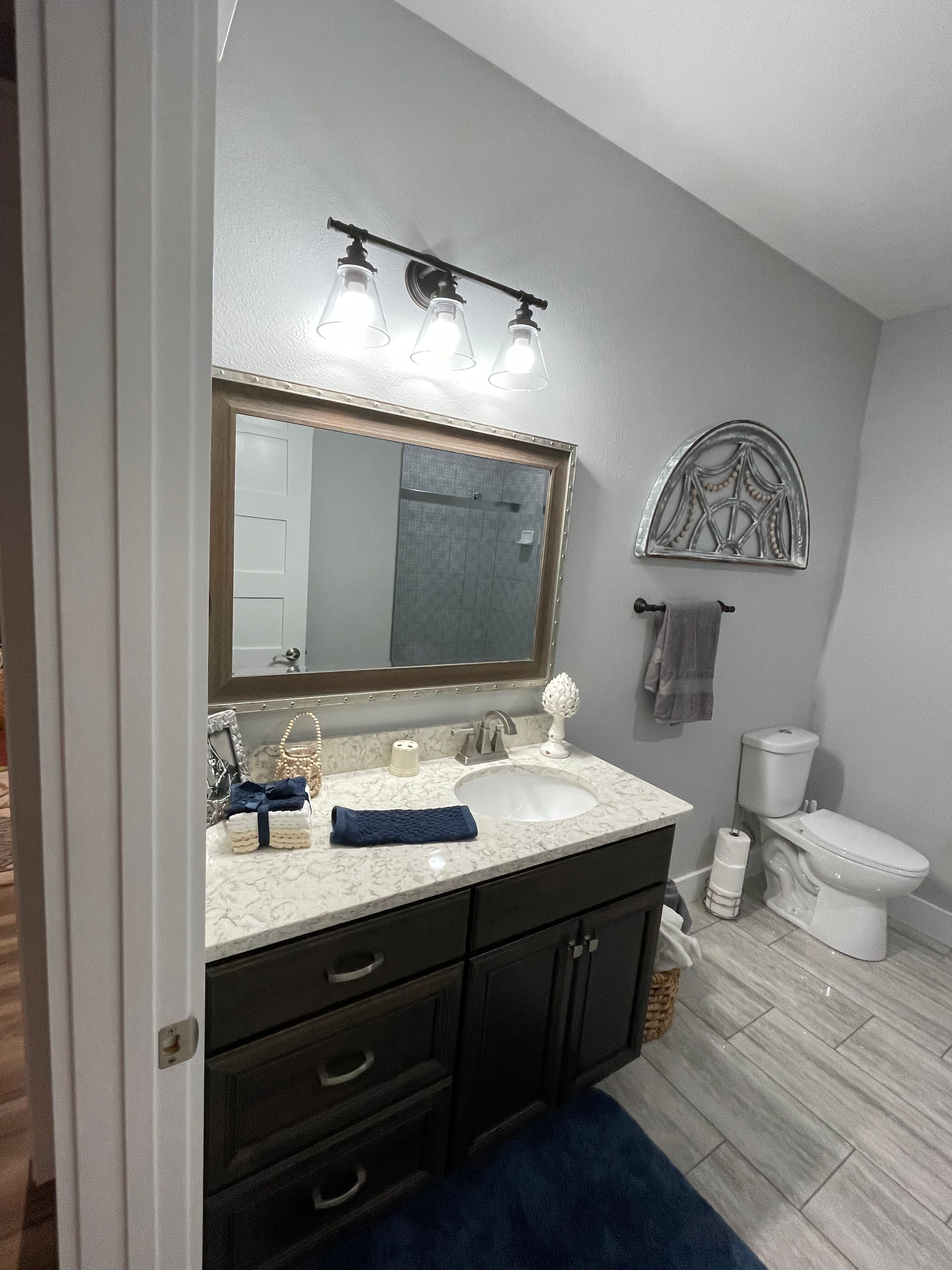 Bathroom with dark vanity, large mirror, sink, wall lights, toilet, and decorative wall shelf.