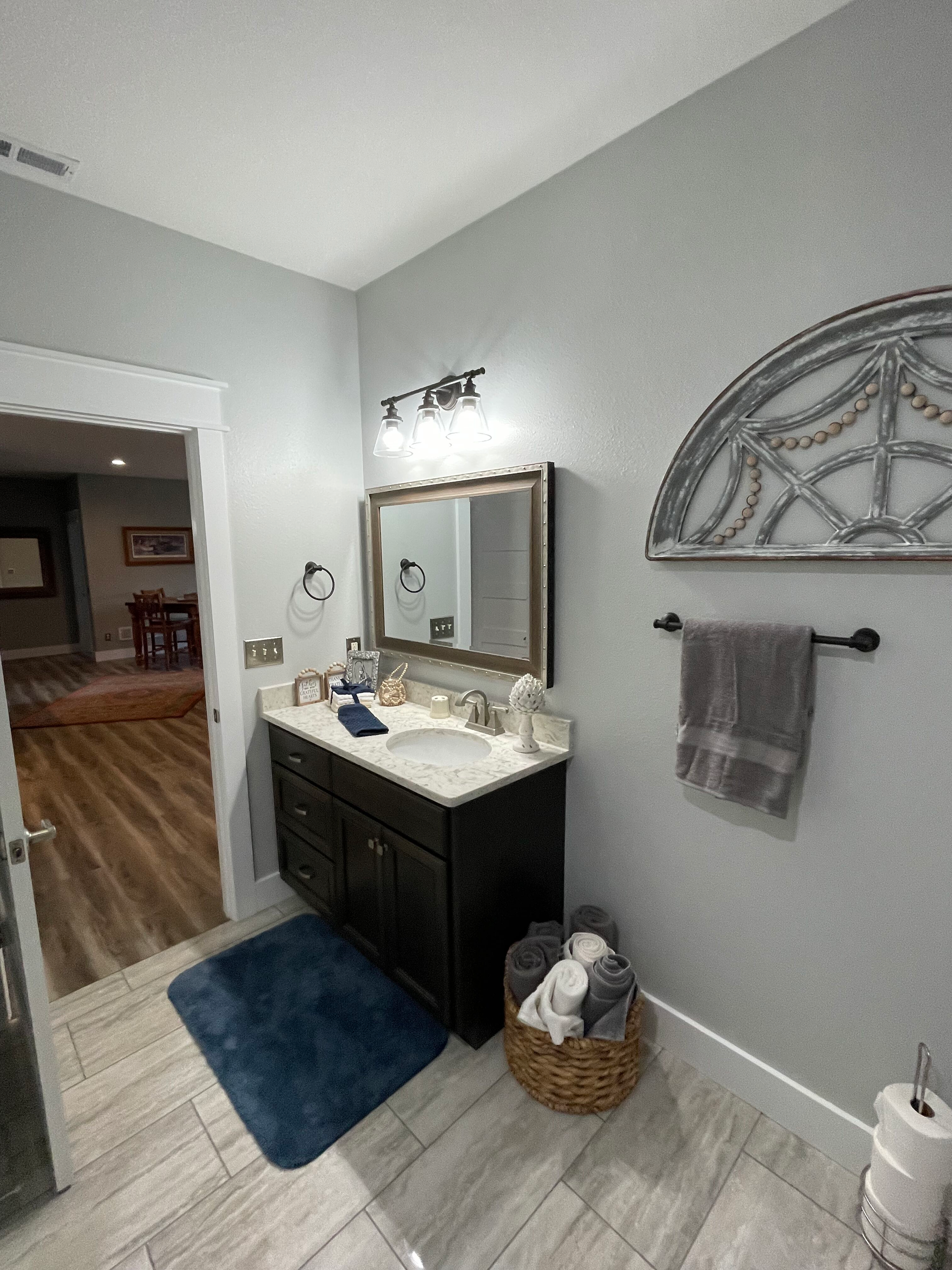 Small bathroom with dark vanity, mirror, blue rug, gray walls, and decorative wall shelf.