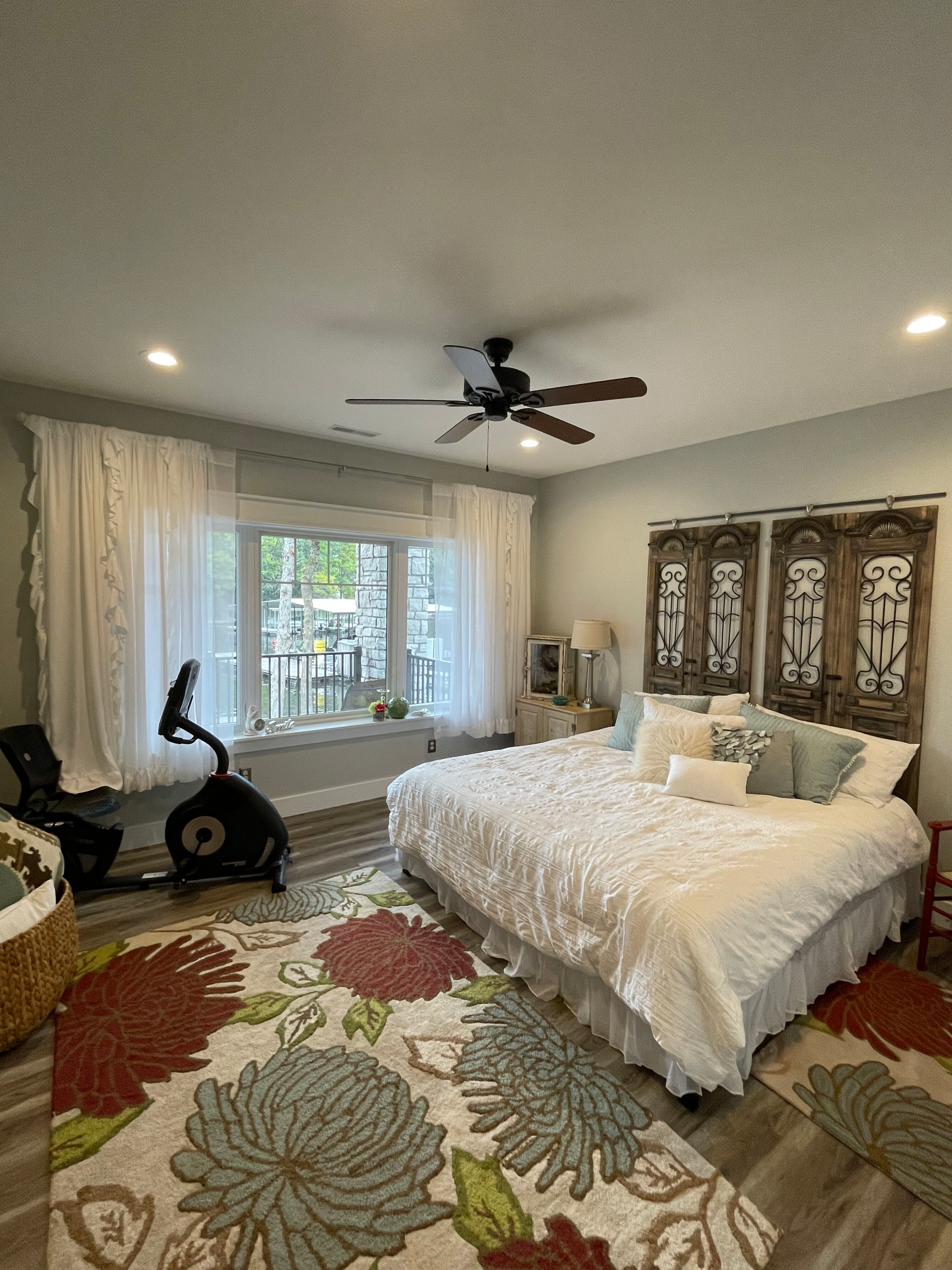 Bright bedroom with floral rug, white bedspread, window seat, exercise bike, and carved wooden headboard