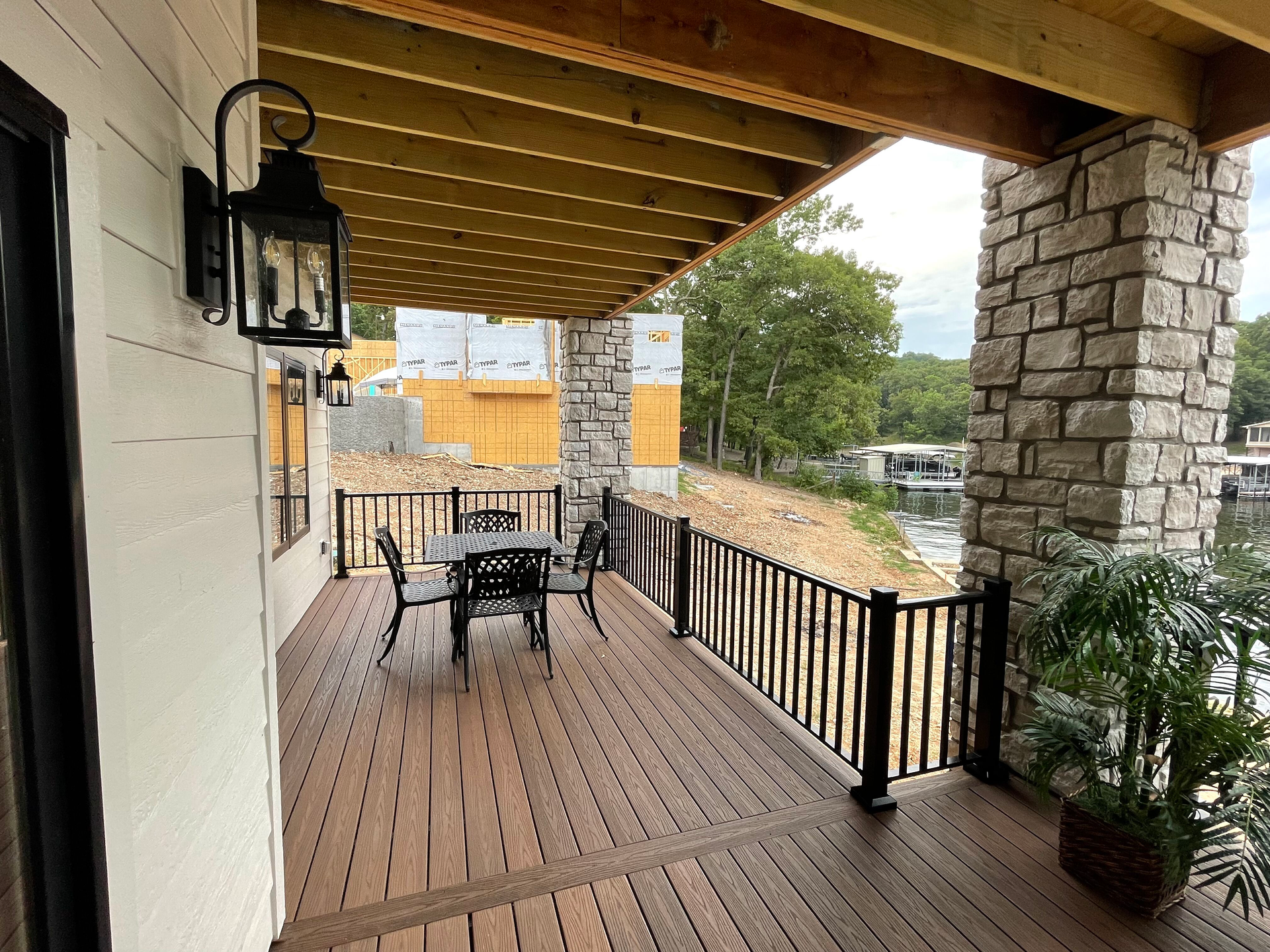 Covered wooden porch with metal patio table and chairs, stone pillar, and black railing overlooking a yard.