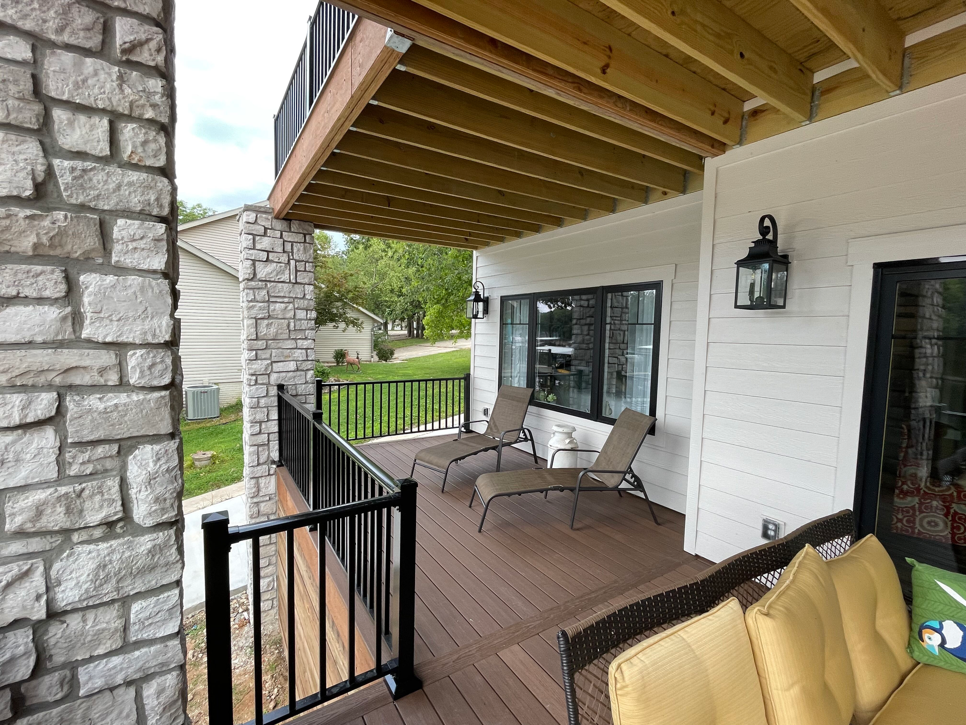 Covered front porch with wooden ceiling, wicker chairs, and black railing beside a stone exterior.