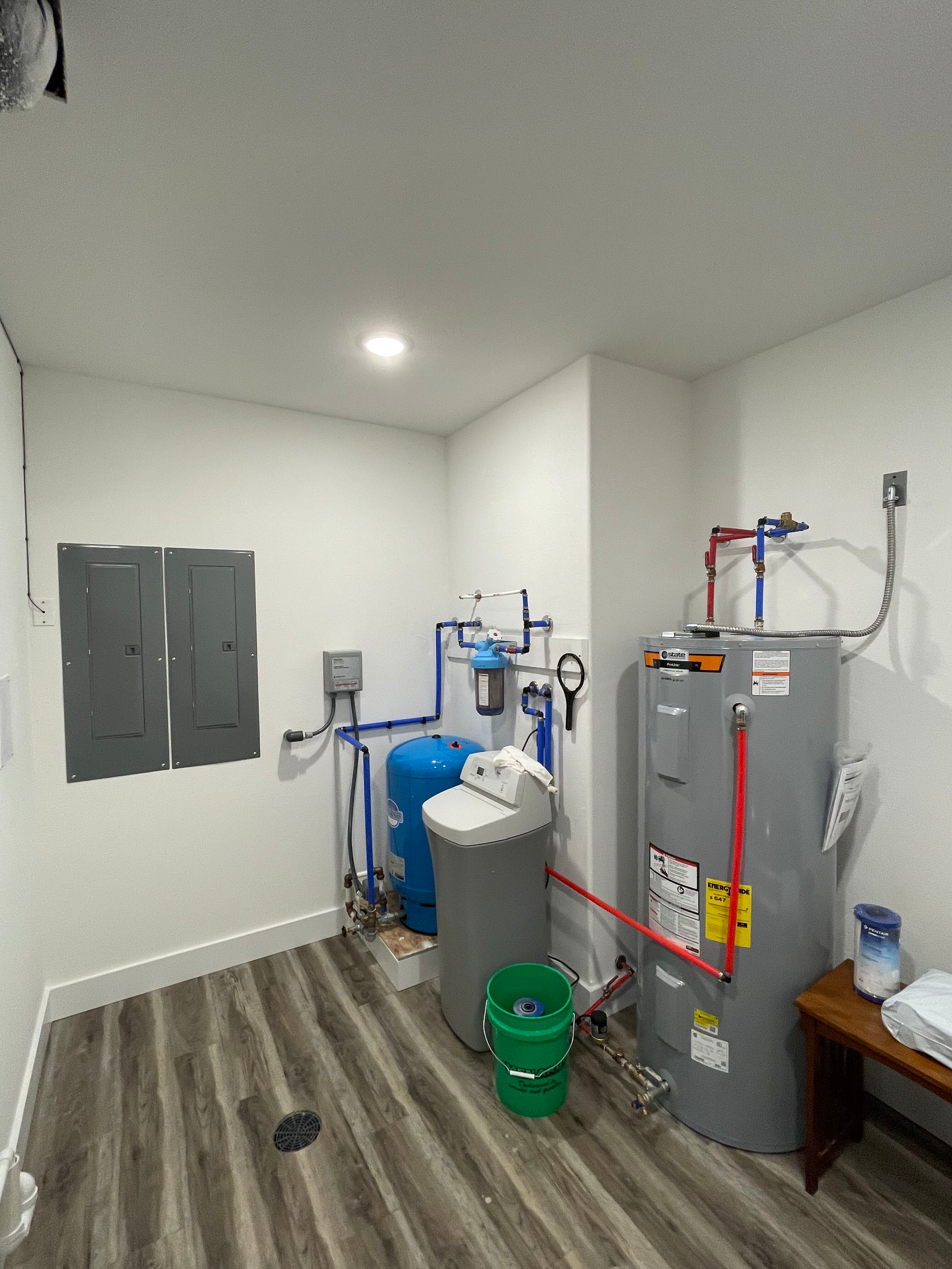 Small utility room with water treatment tanks, gray electrical panel, and plumbing on wood-look floor.