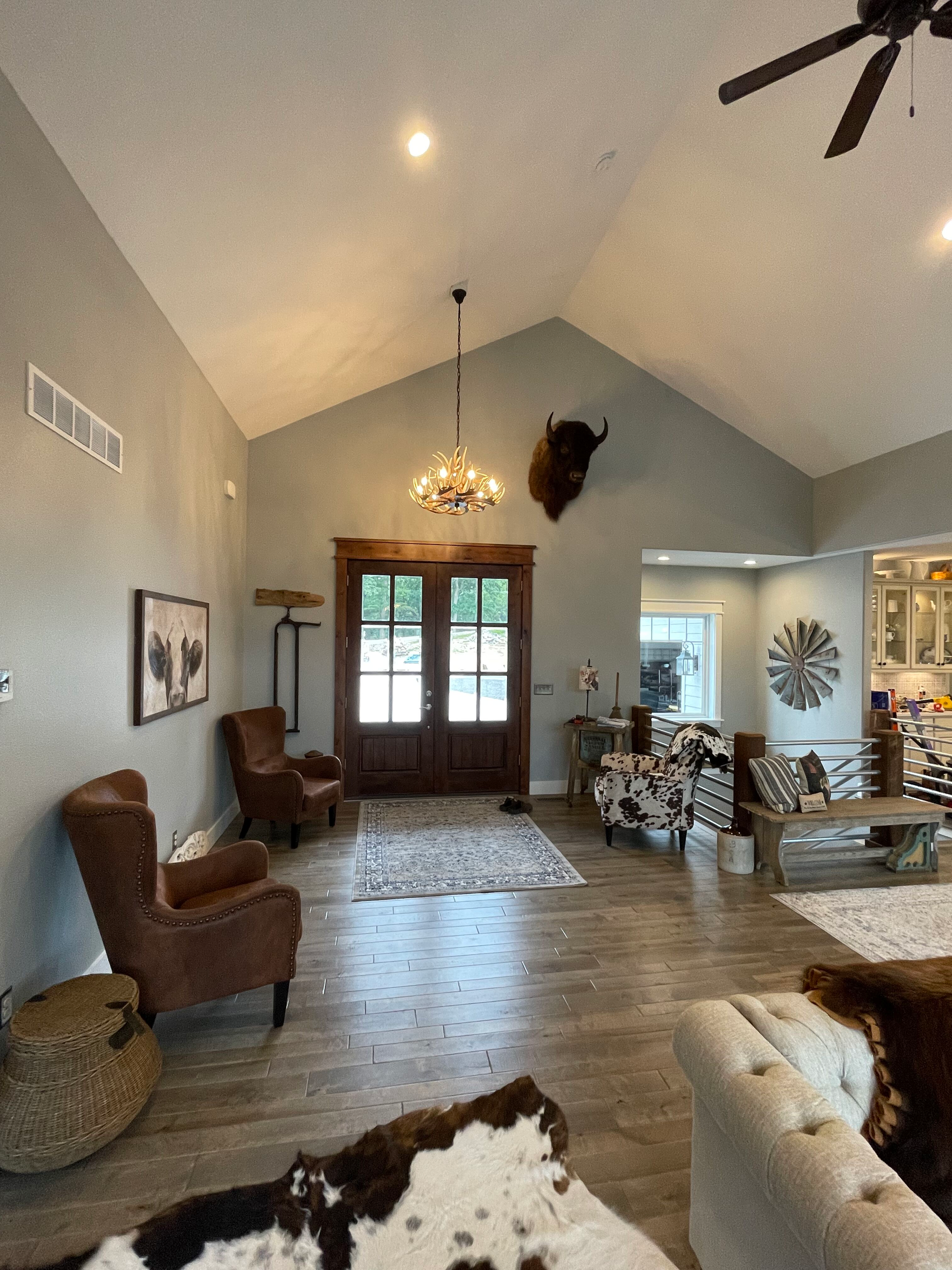 Living room with vaulted ceiling, chandelier, brown chairs, patterned rug, and French doors by the fireplace.