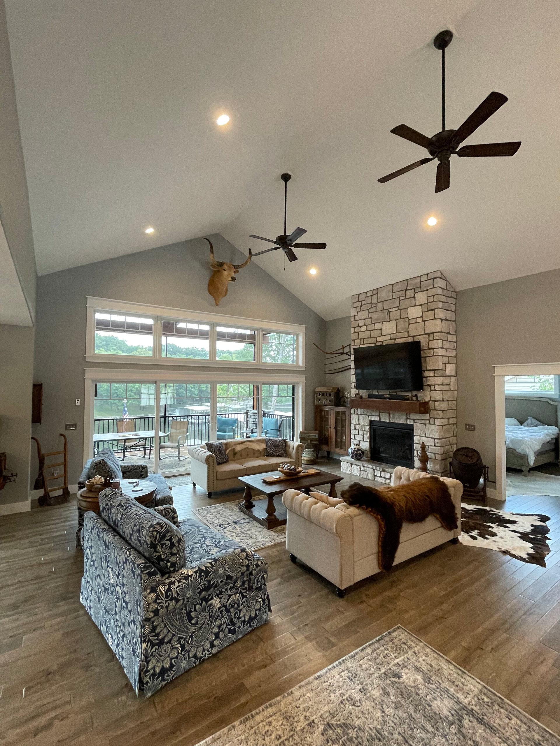 Bright living room with stone fireplace, large windows, wood floors, and modern ceiling fans.