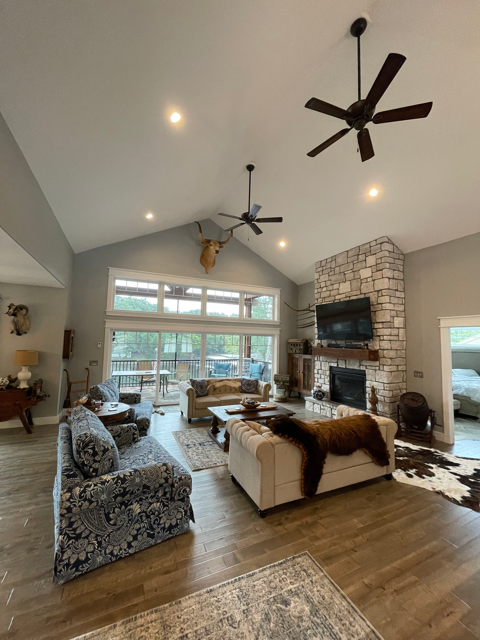 Spacious living room with vaulted ceiling, stone fireplace, patterned sofa, and large windows opening to greenery