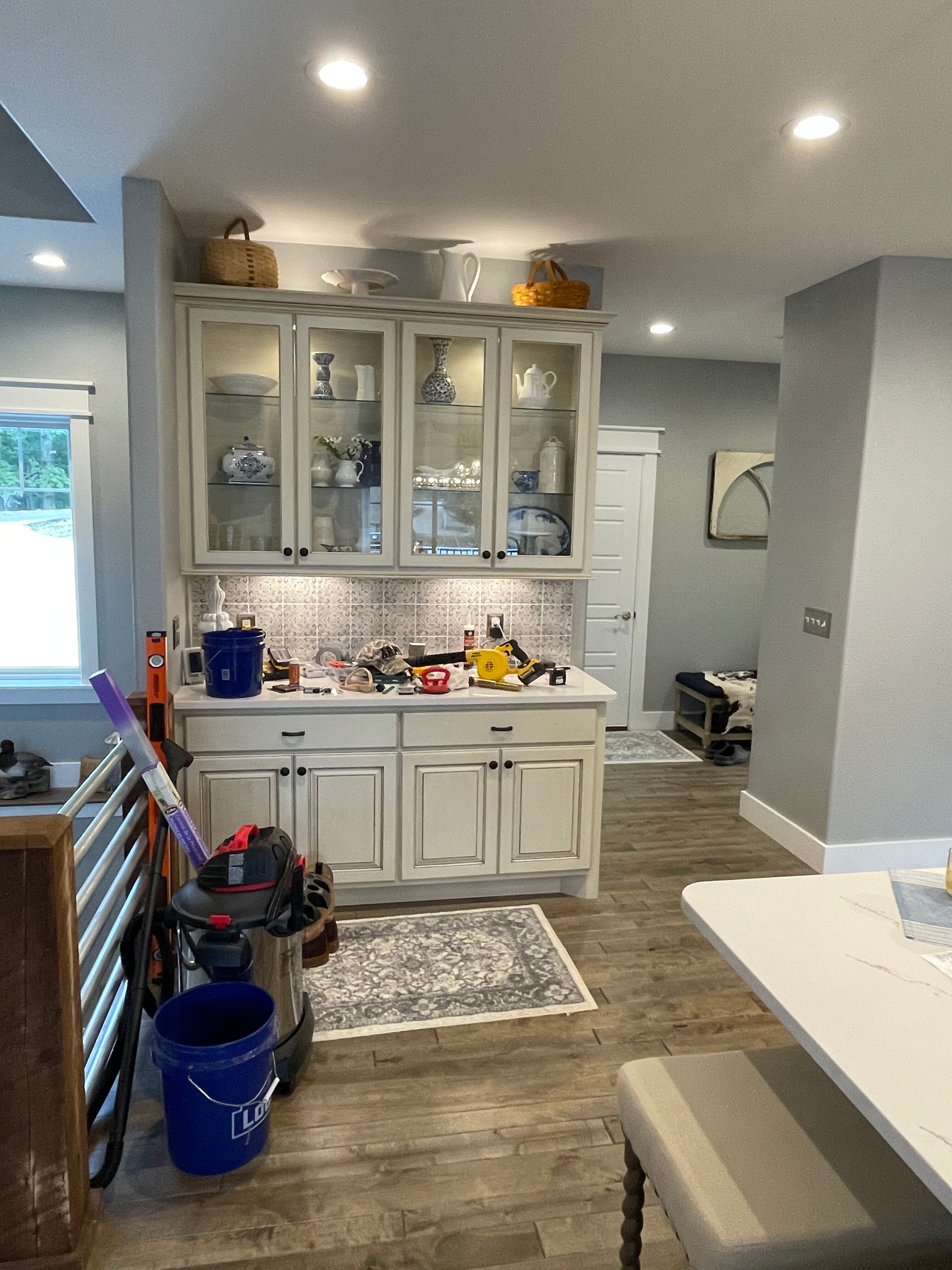 Messy kitchen with white cabinets, wood floors, cleaning supplies, and a blue bucket in the foreground