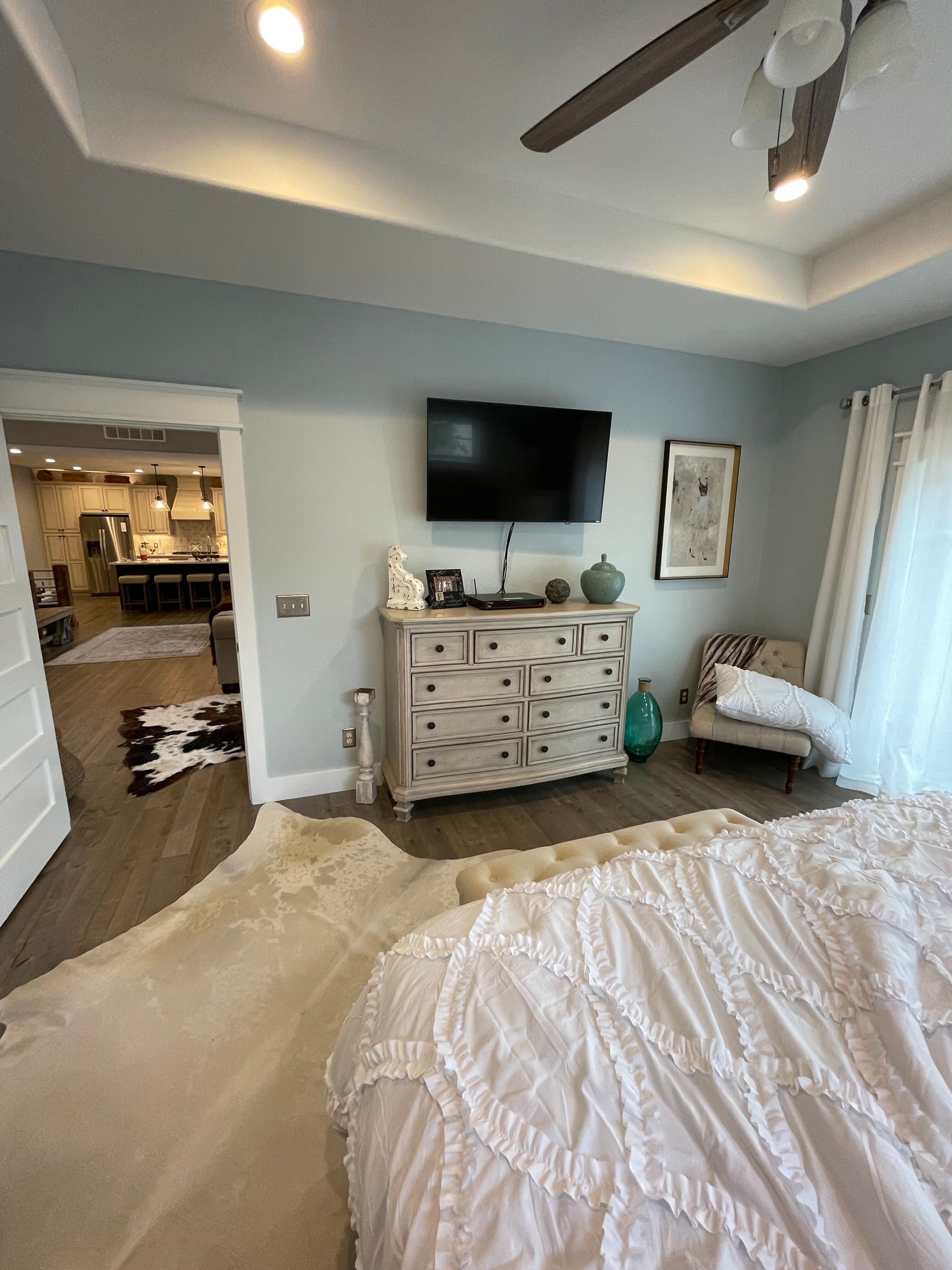Bedroom with white bedspread, dresser with TV, ceiling fan, and open doorway to a lit adjoining room.