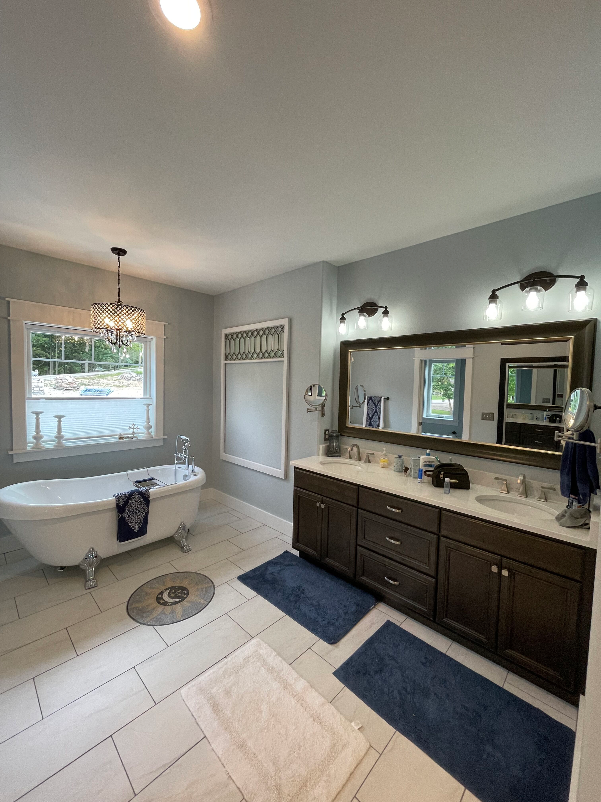 Bright bathroom with clawfoot tub, dark vanity, large mirror, blue rugs, and window light