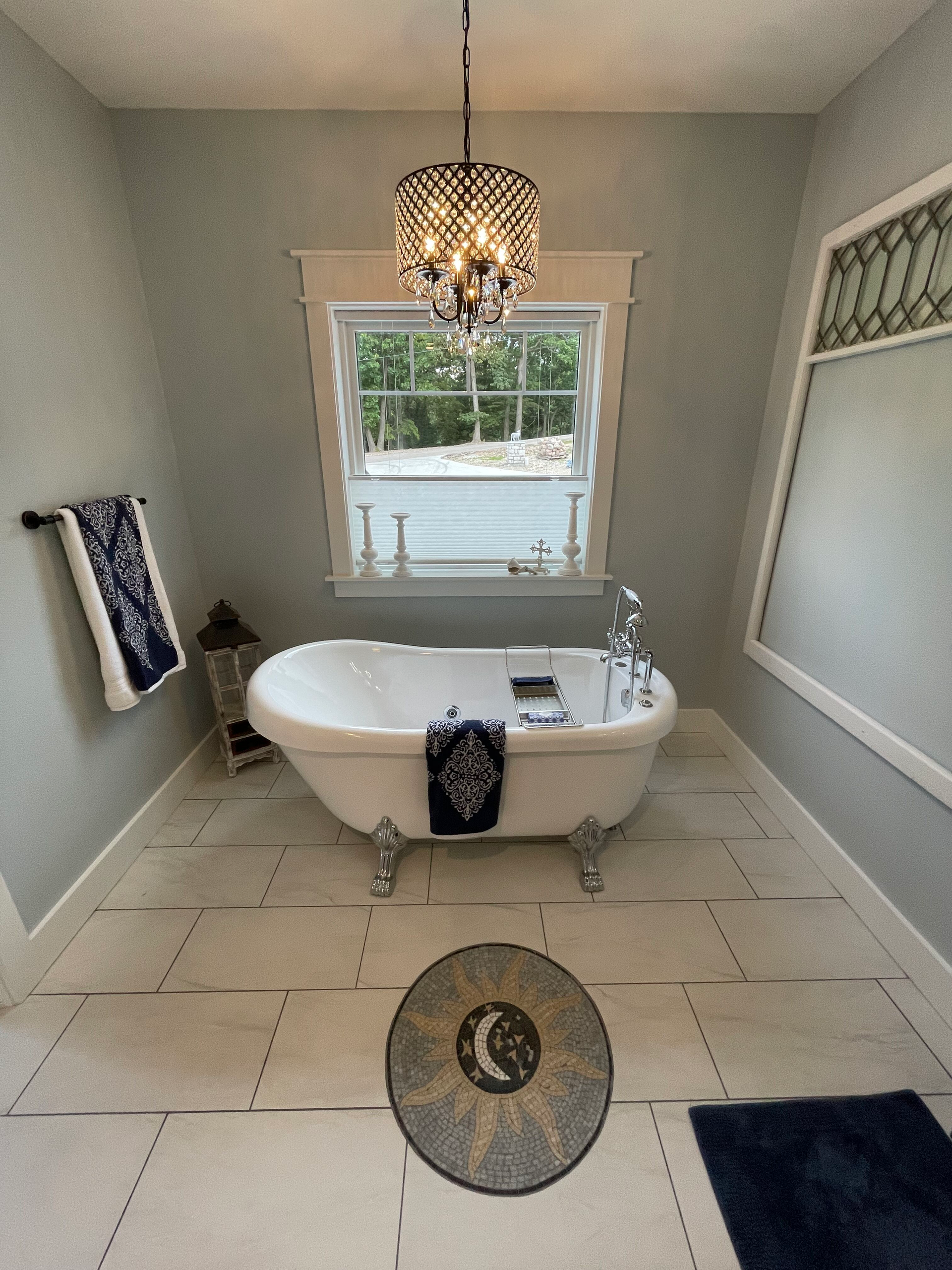 Bright bathroom with a clawfoot tub, chandelier, large window, and beige tile floor.