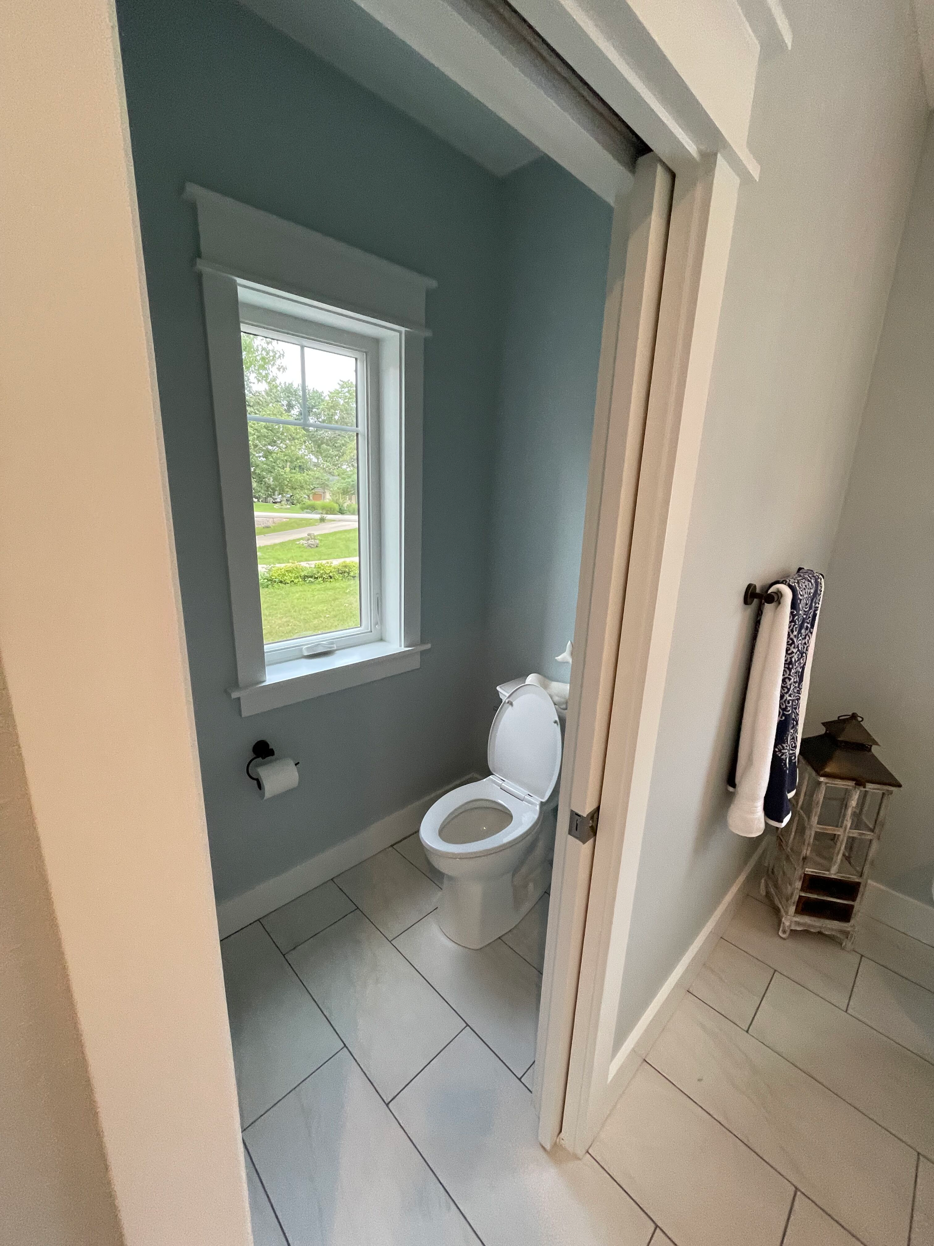 Small bathroom with a toilet, window, and light gray tile floor, viewed through an open doorway