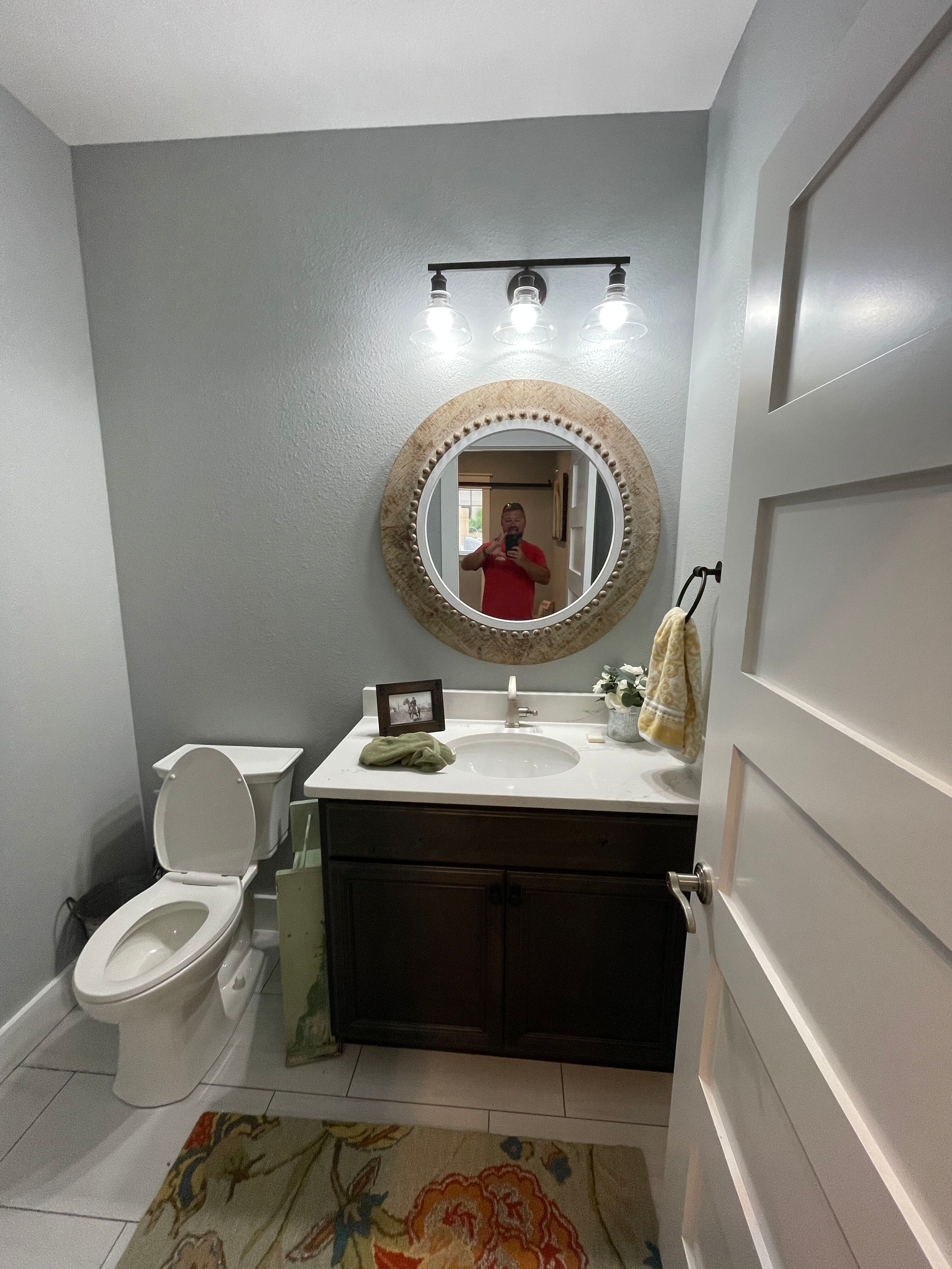Small bathroom with toilet, dark vanity, round mirror, and a person reflected in the mirror.