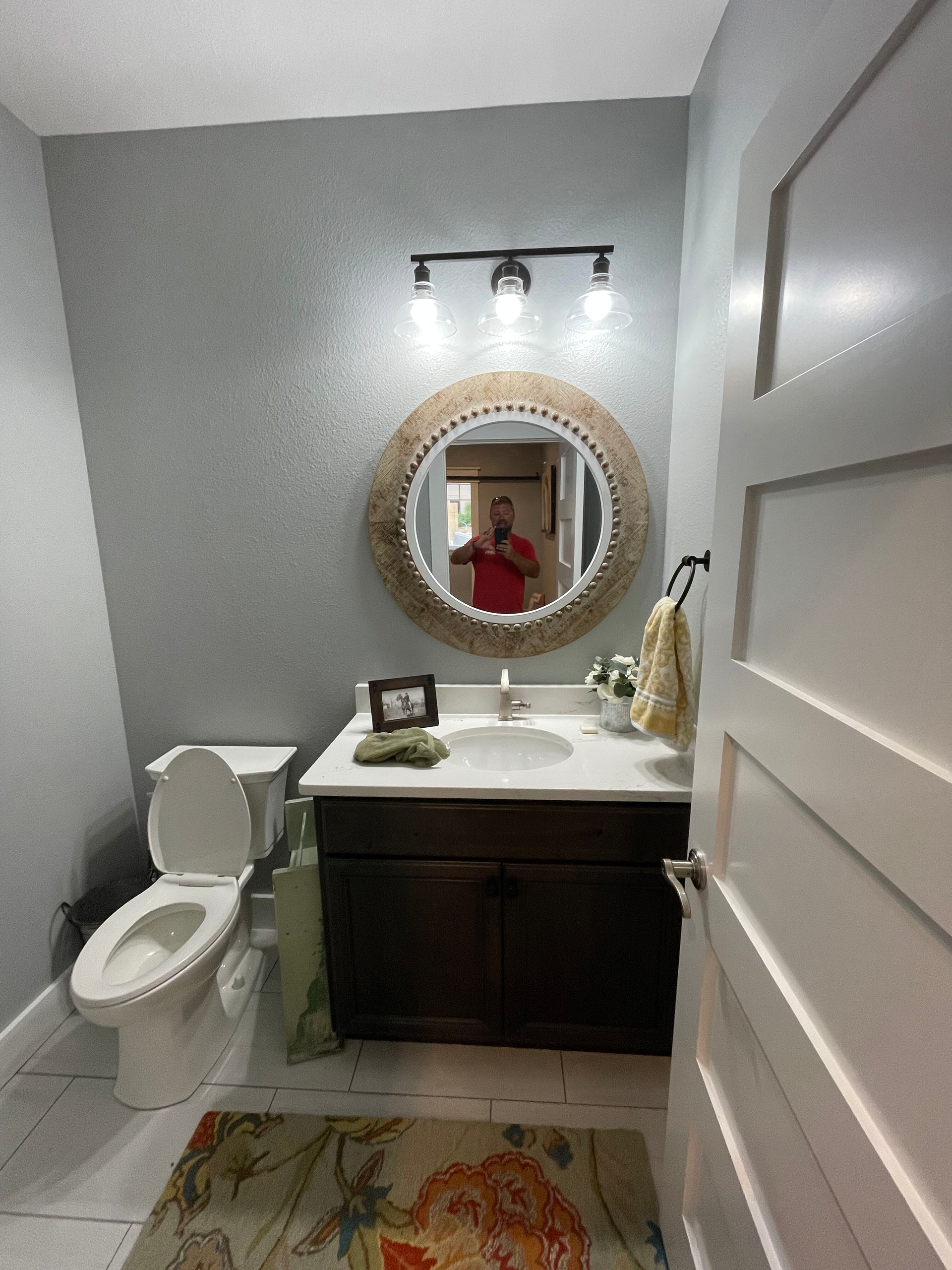 Small bathroom with toilet, dark vanity, round mirror, wall sconce, and floral rug.