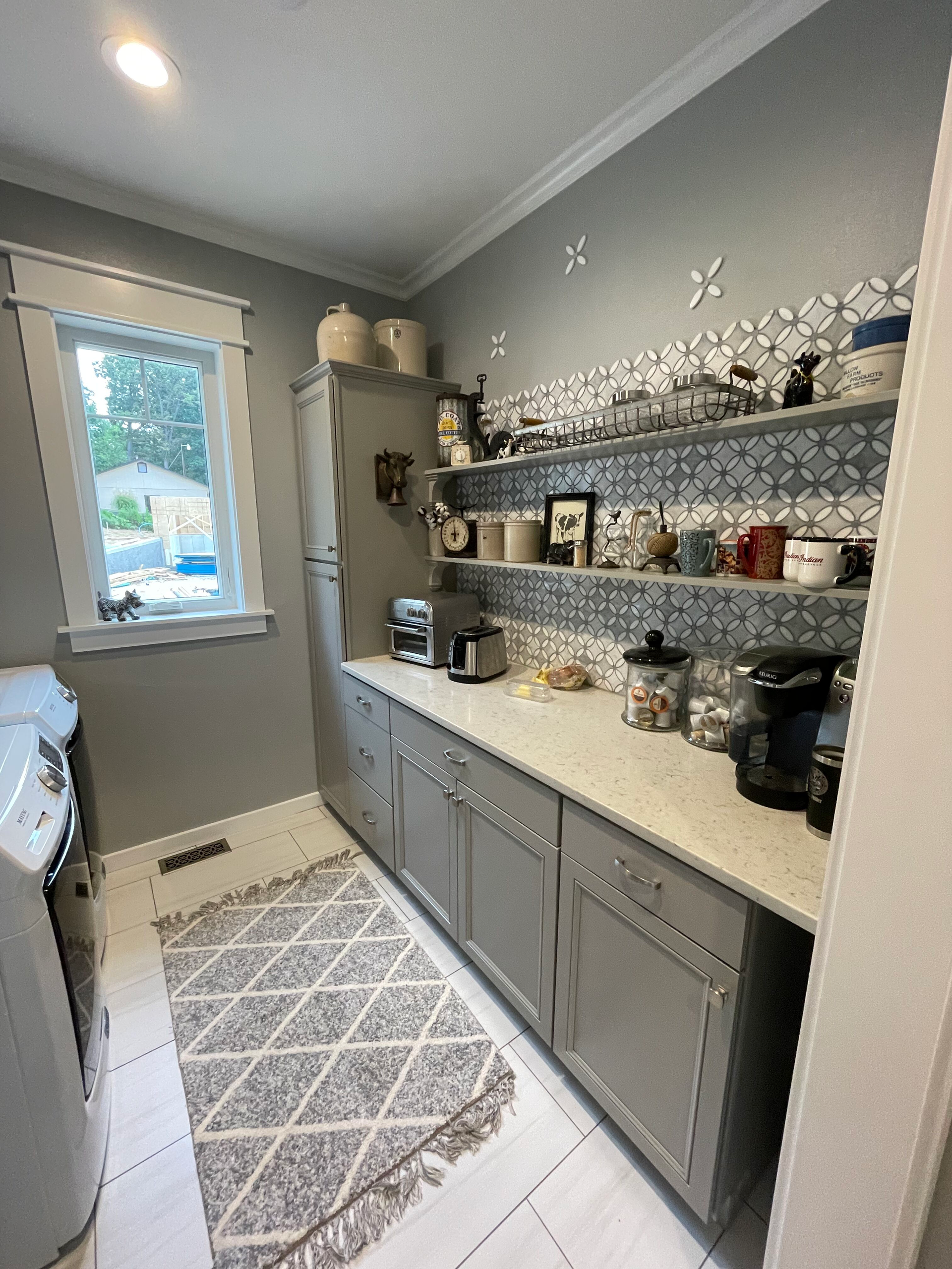 Narrow gray kitchen with open shelves, countertop appliances, and a patterned rug by a window.