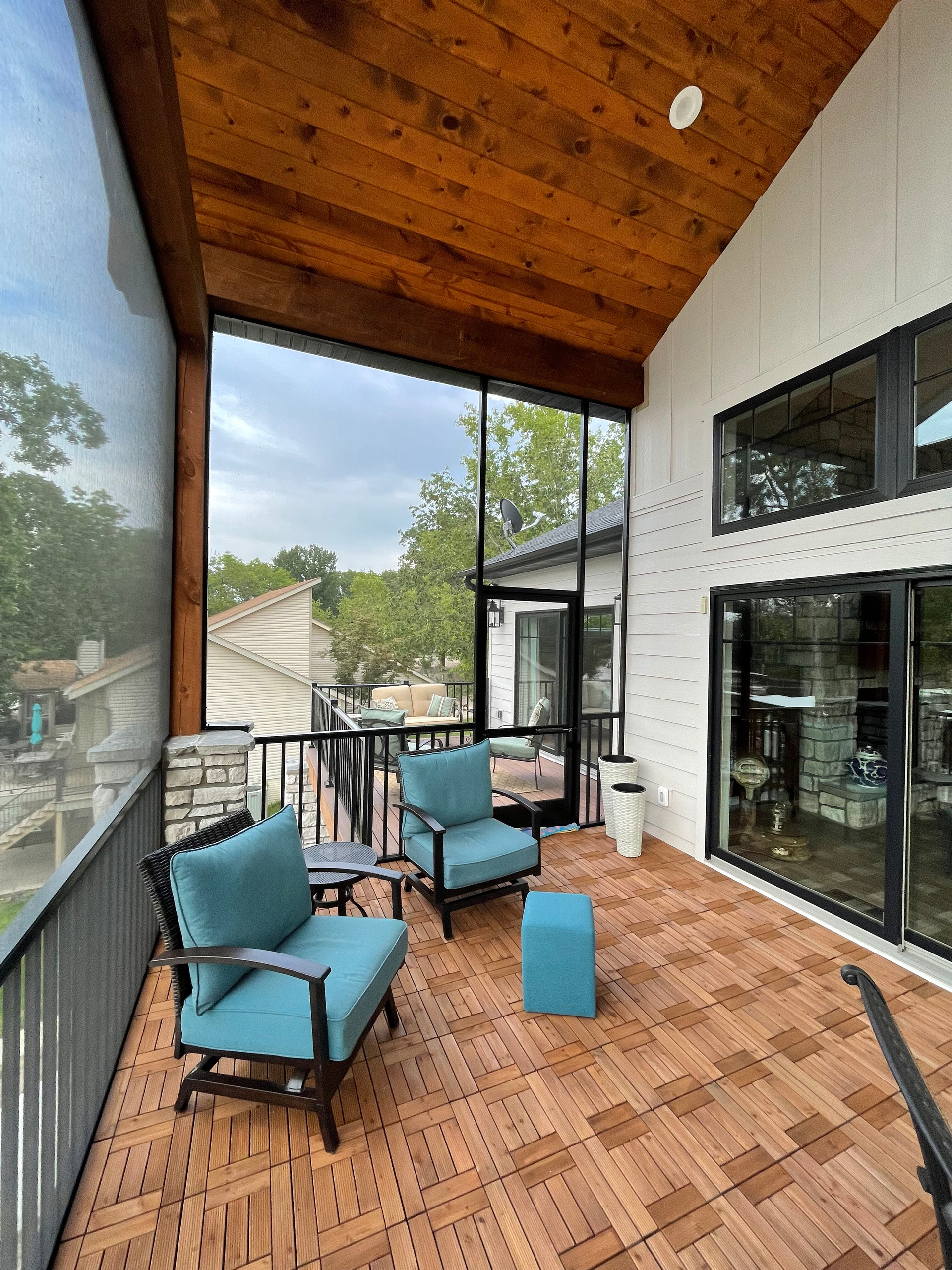 Covered balcony with blue cushioned chairs, small table, and wood ceiling overlooking a tree-lined view.