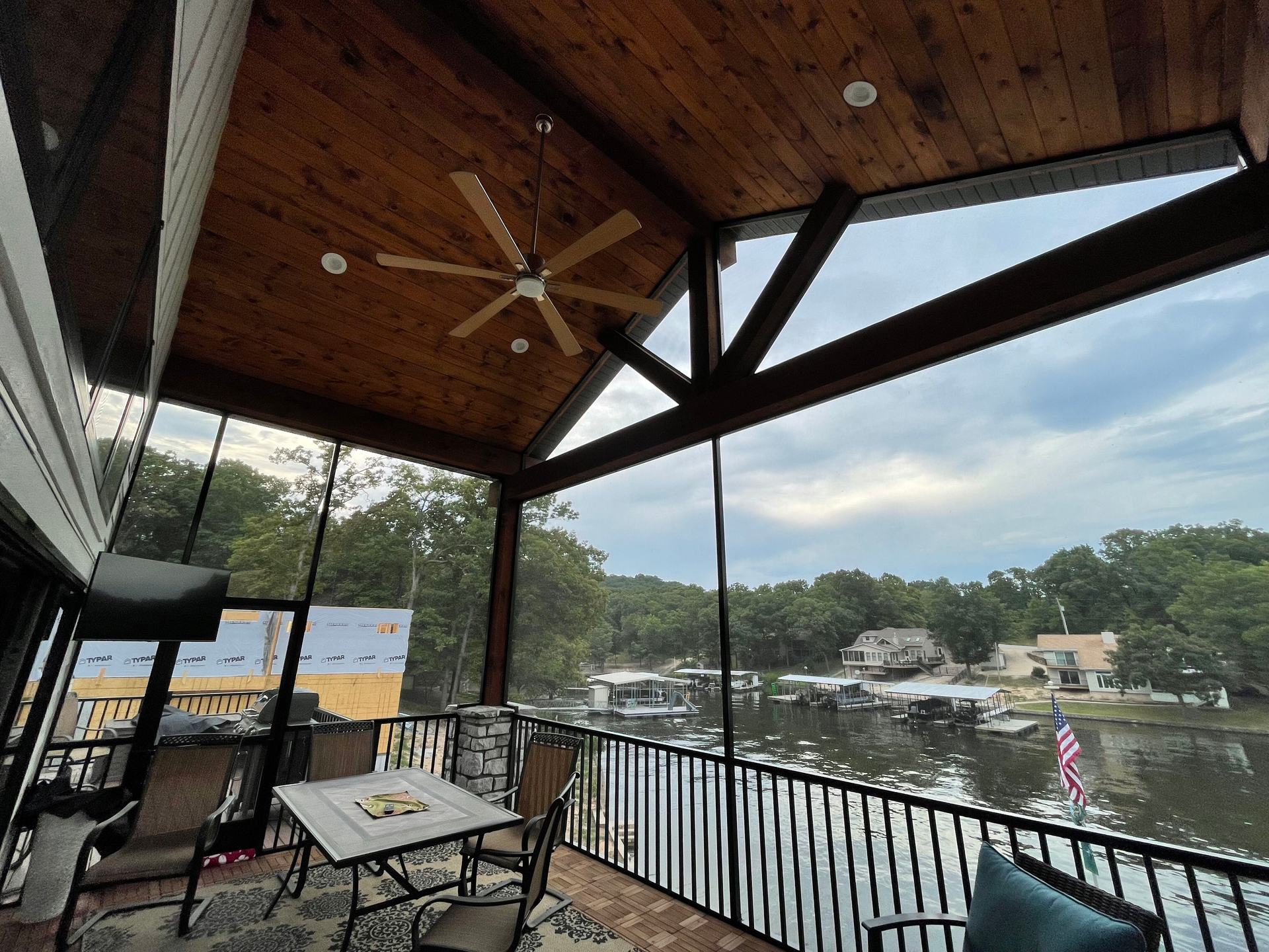 Covered lakeside deck with wooden ceiling, railing, and tables overlooking a calm lake and green hills.