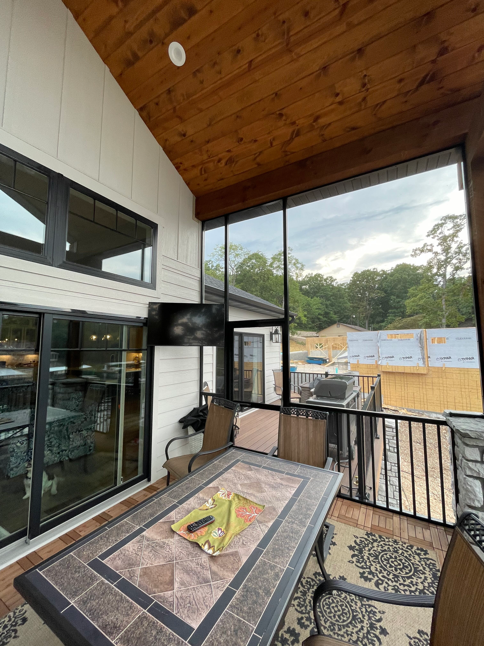 Covered balcony with black railings, glass doors, tiled table, and wooded view beyond construction site.
