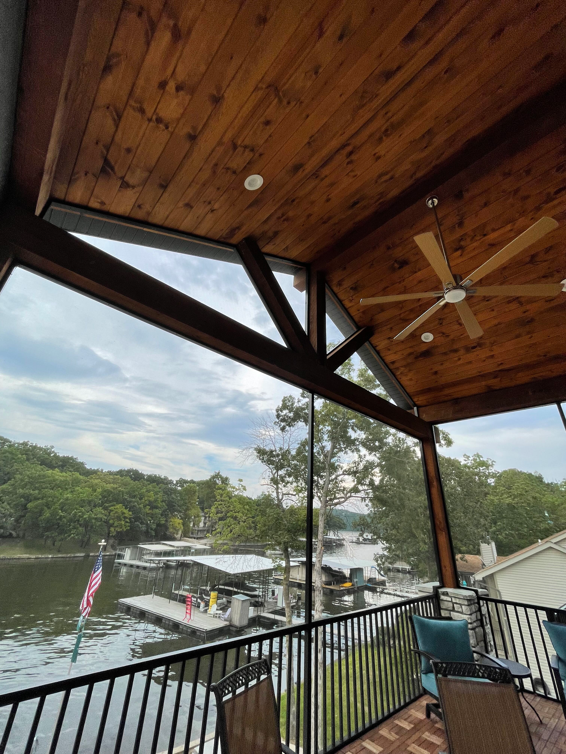 Covered balcony overlooking a river, boats, and trees, with wooden ceiling beams and a ceiling fan