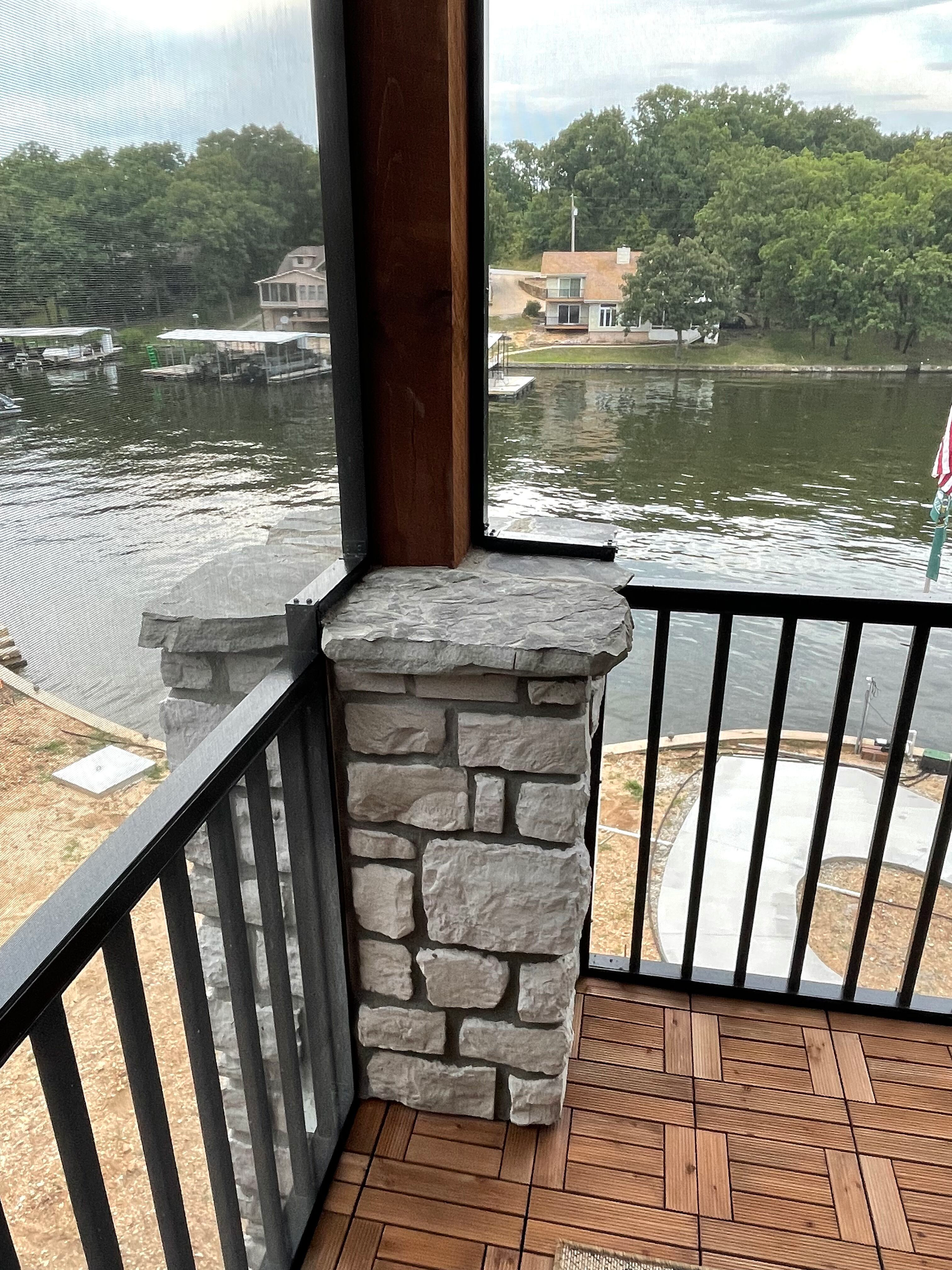 Stone porch overlooking a lake with black railing and white chairs, viewed through a window.