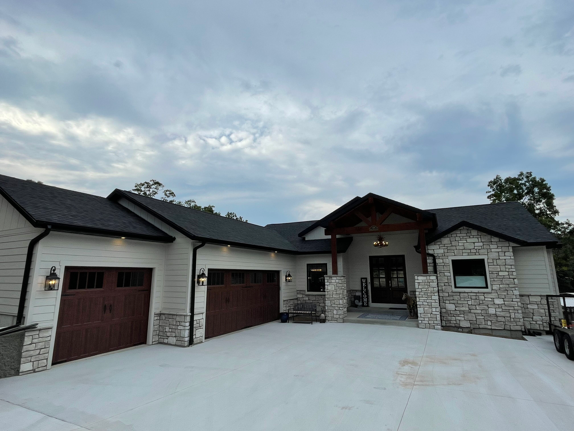 Modern stone house with dark roof, two garages, and a large concrete driveway under a cloudy sky