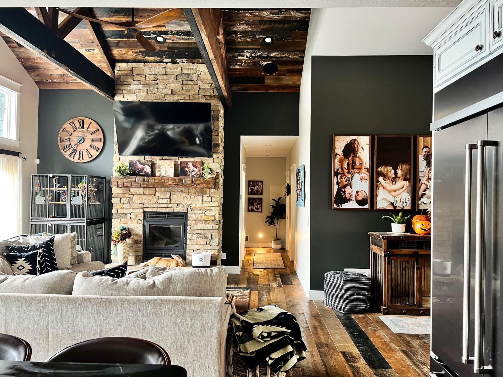Cozy living room with exposed wood beams, stone fireplace, TV, and stainless steel fridge in open kitchen