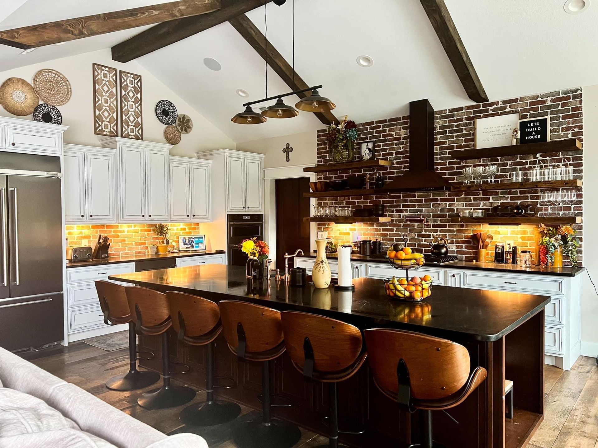 Modern kitchen with dark island, bar stools, white cabinets, exposed beams, and brick backsplash.