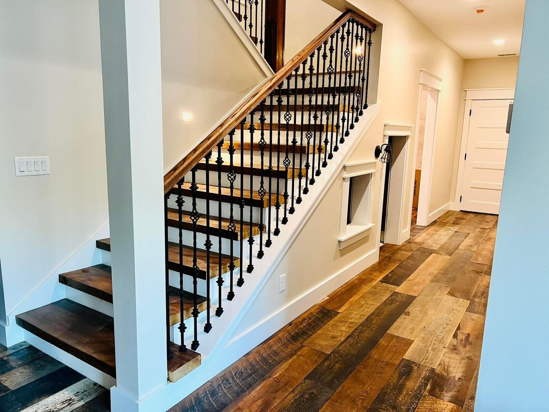 Staircase in a bright hallway with black balusters, white walls, and wood plank flooring