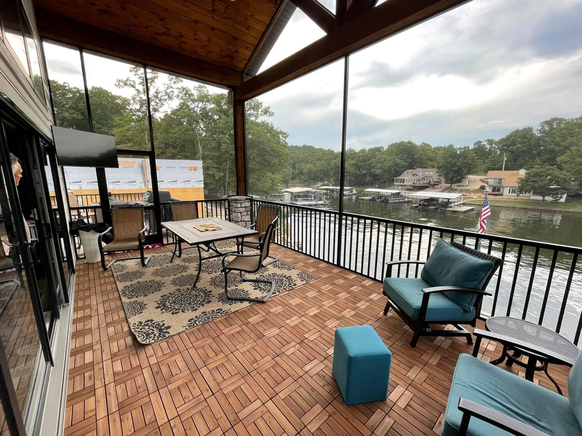 Covered riverside patio with wicker seating, patterned rug, and lake view under a wooden roof