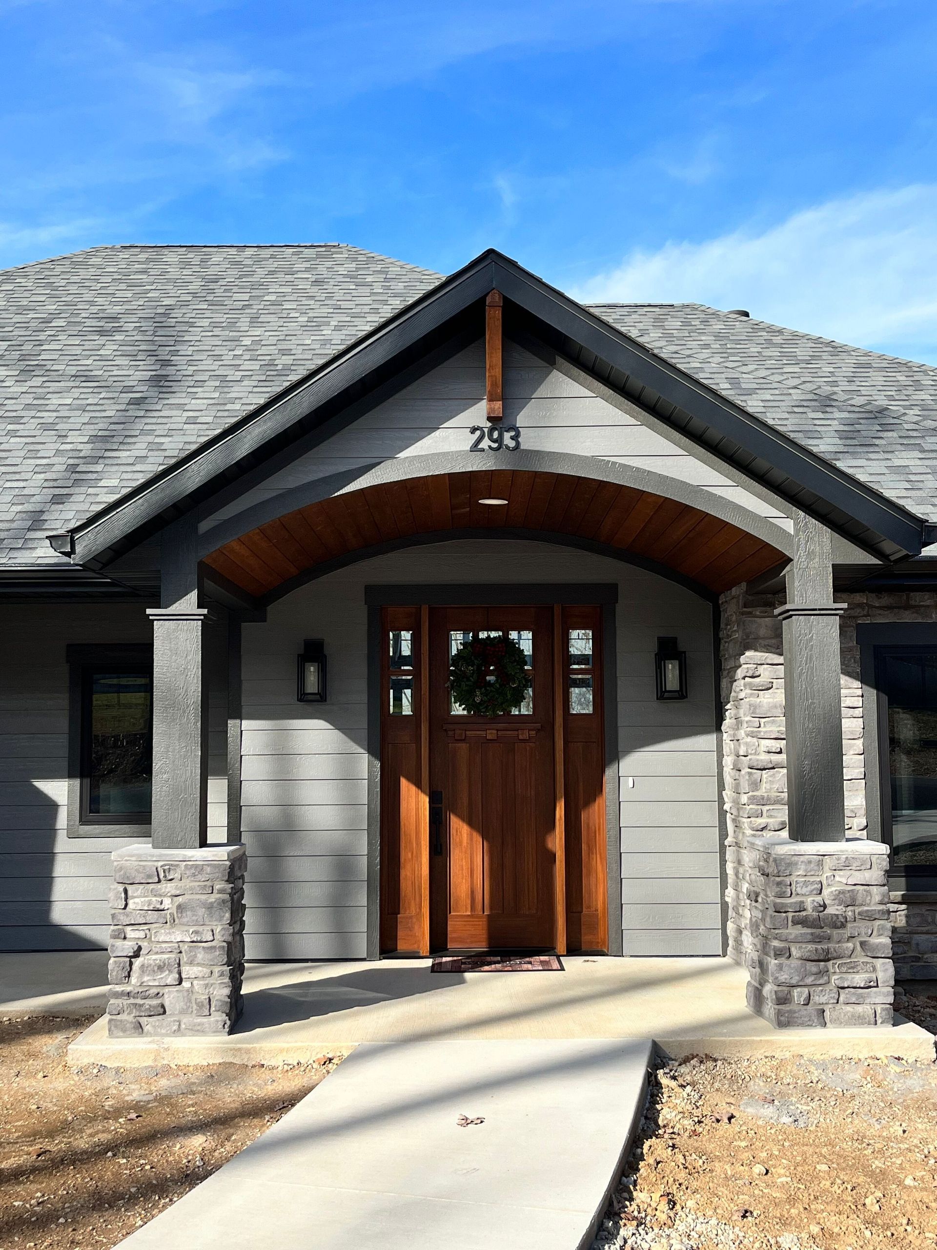 Front entrance of a stone house with wooden double doors, covered porch, and concrete walkway under a blue sky
