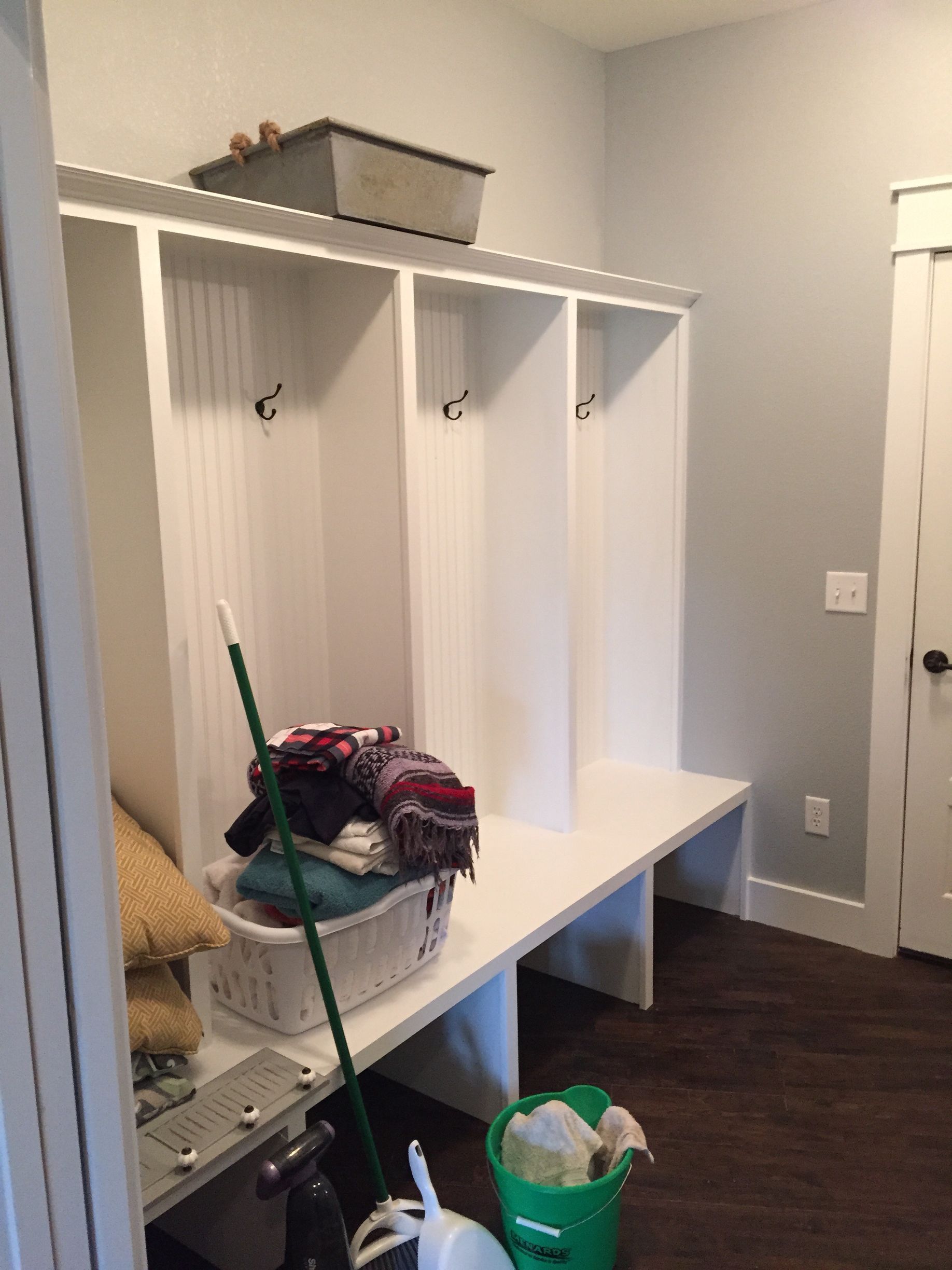 White mudroom bench with cubbies and hooks, holding cleaning supplies and bags, next to a door.