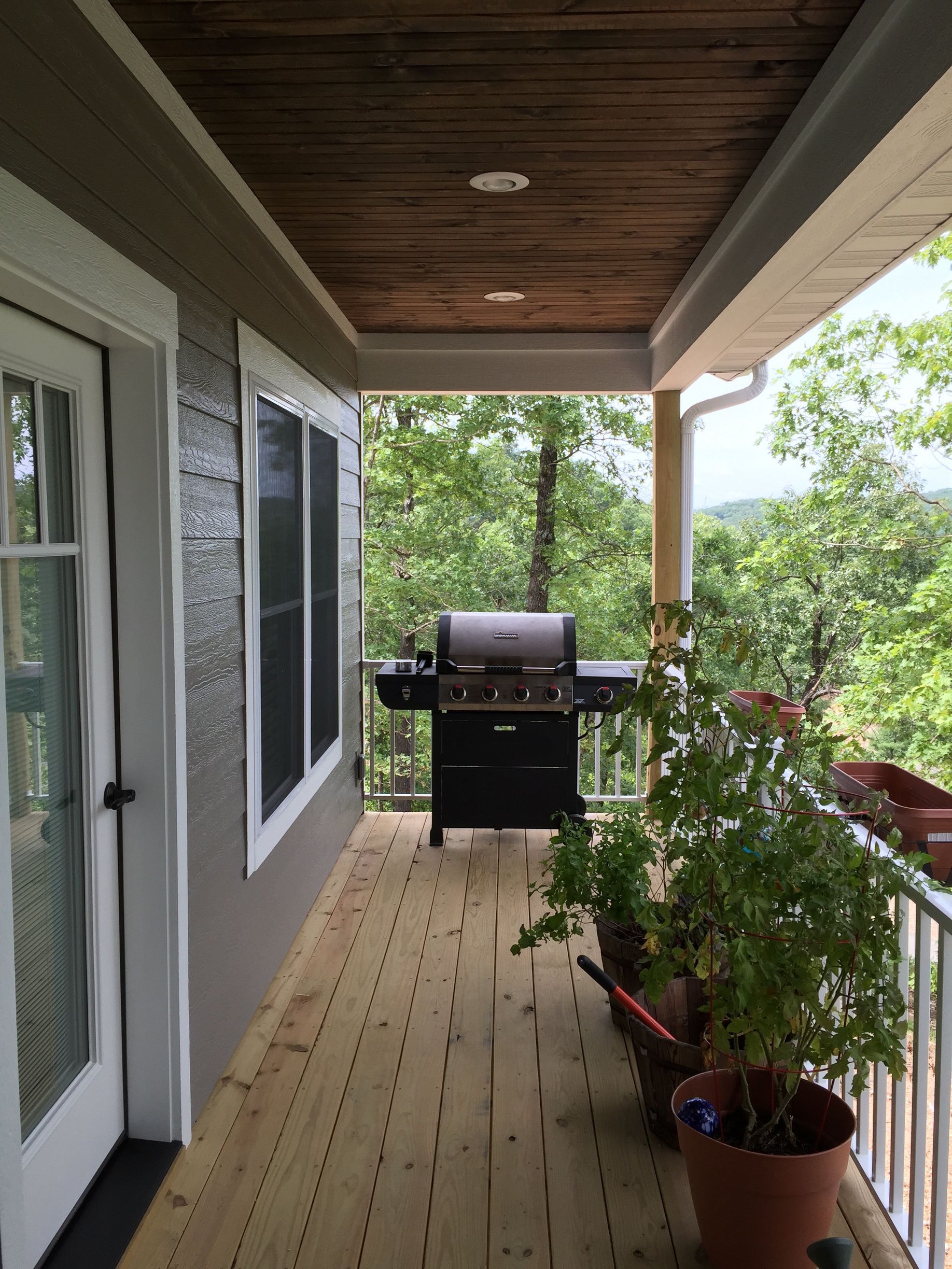 Covered porch with wooden floor, grill, potted plants, and view of trees outside.