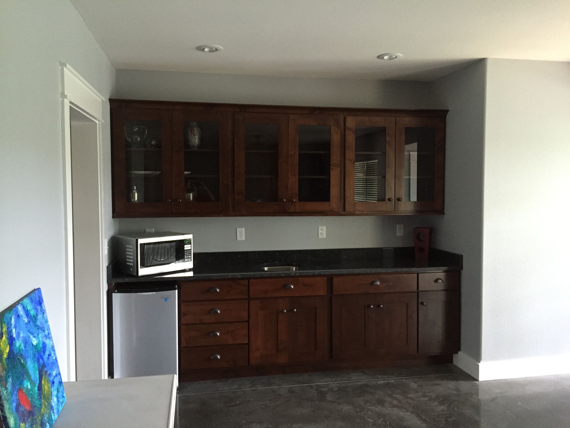 Kitchen with dark wood cabinets, black countertop, mini fridge, and microwave against a light wall.