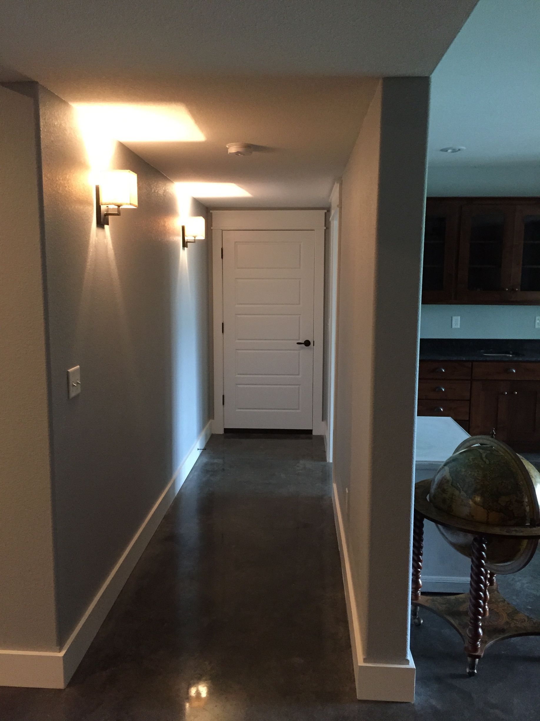 Long hallway with dark floors, gray walls, and a white door at the end, lit by a wall sconce
