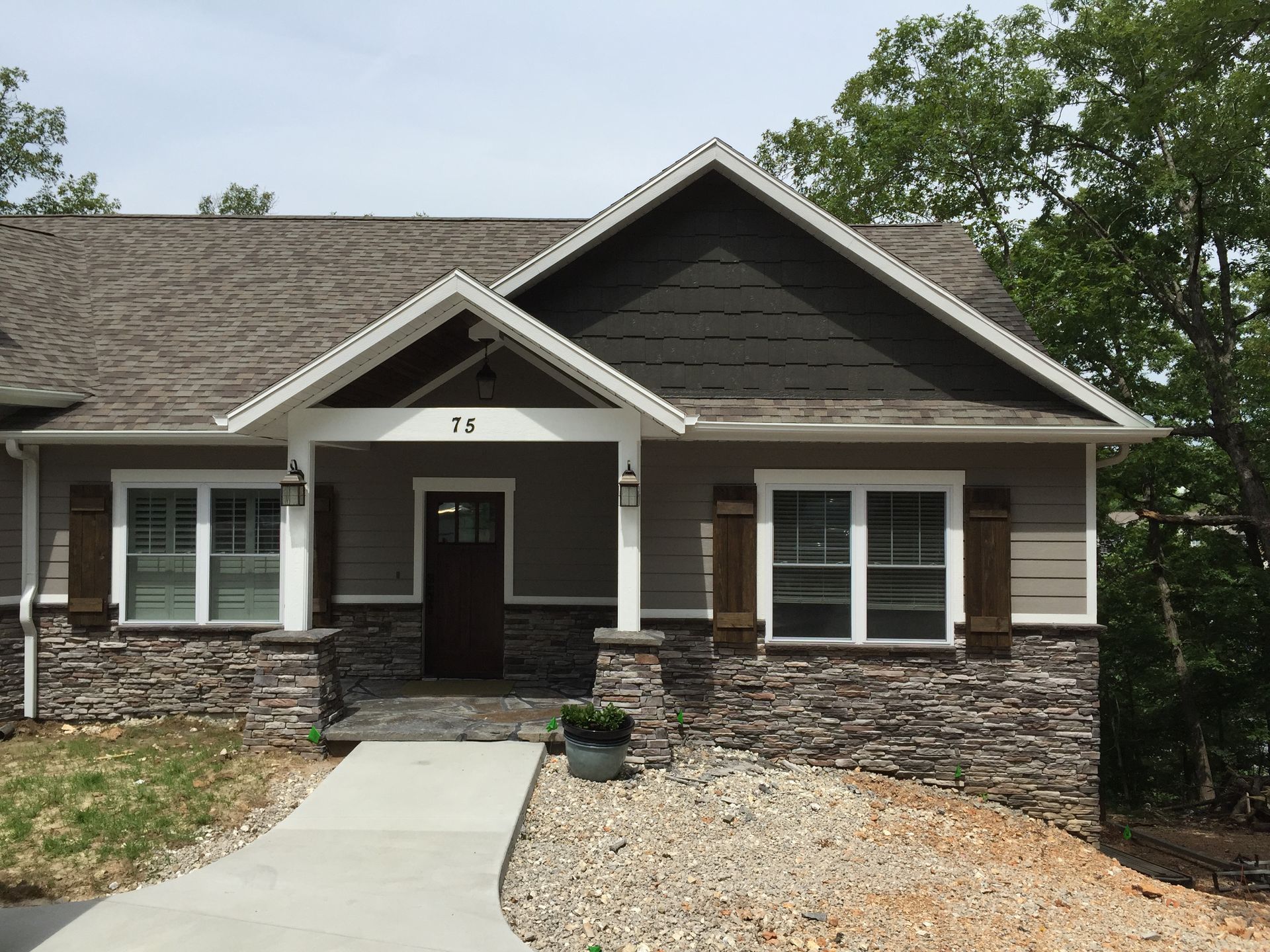 Single-story house with dark siding, stone base, and a front walkway in a wooded setting