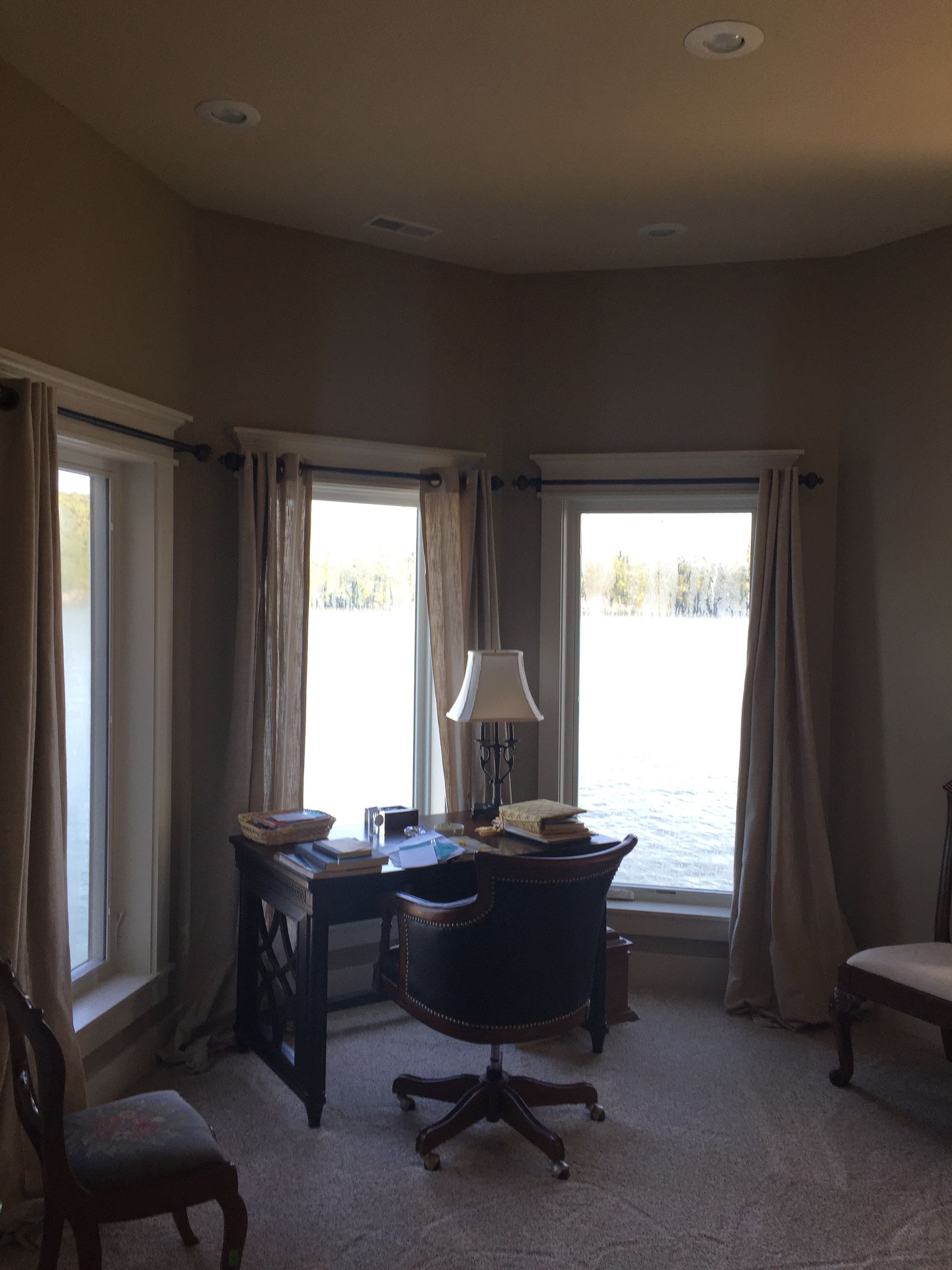 Home office with desk and lamp in front of bright bay windows, curtains, and upholstered chairs