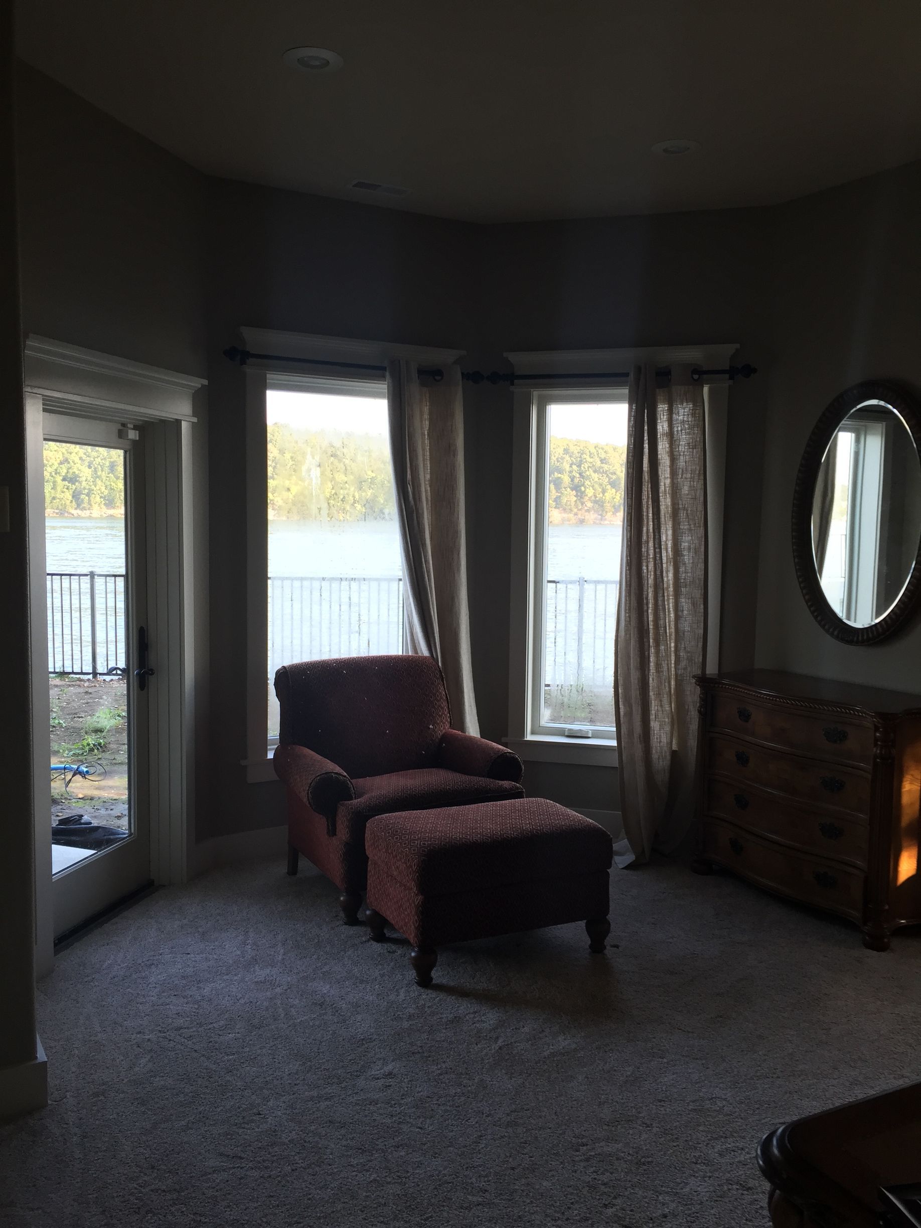 Dim living room with two chairs, large windows, gray carpet, and a wooden dresser with oval mirror.