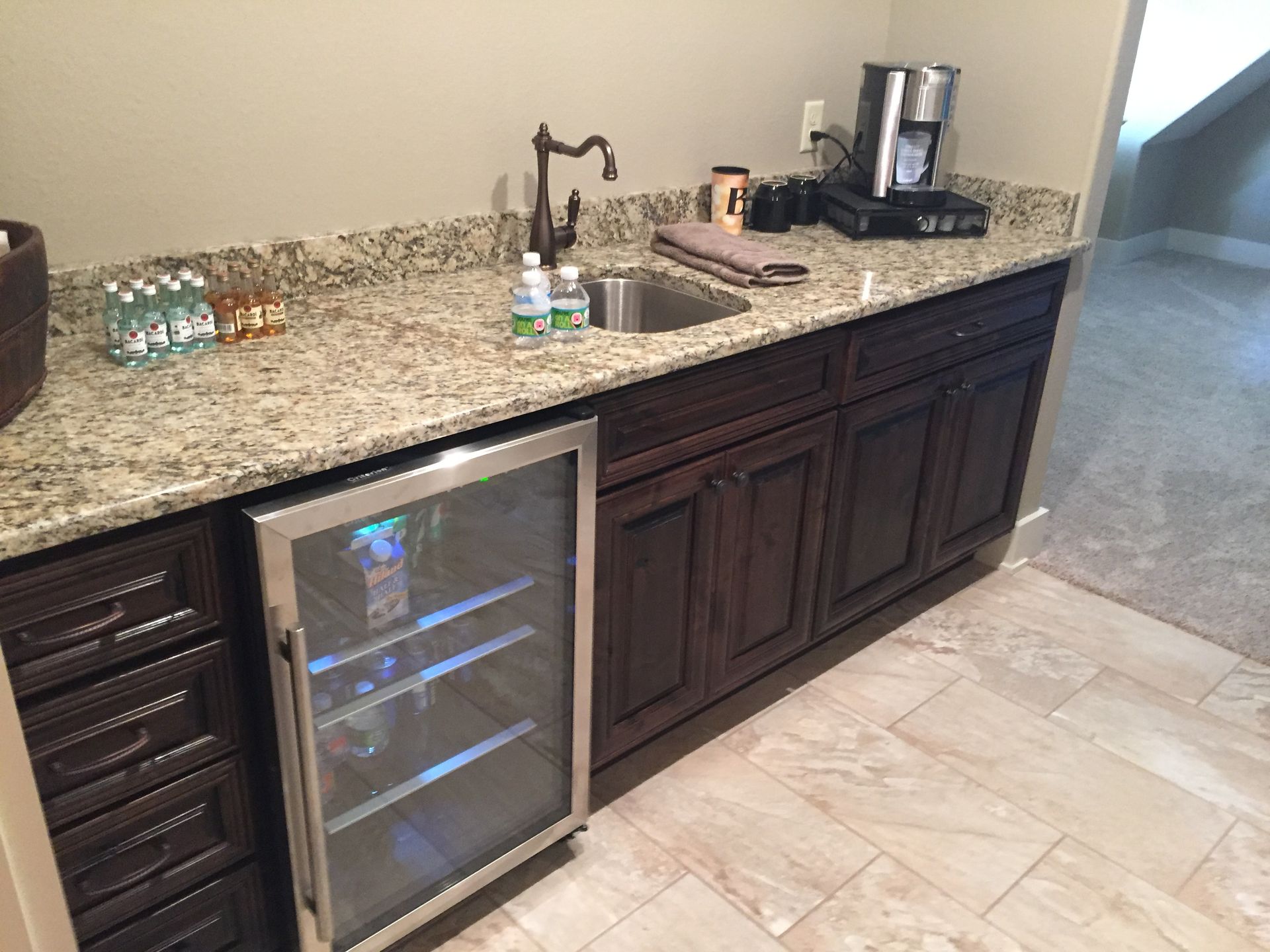 Granite kitchen counter with wine fridge, coffee maker, and dark wood cabinets next to tiled floor
