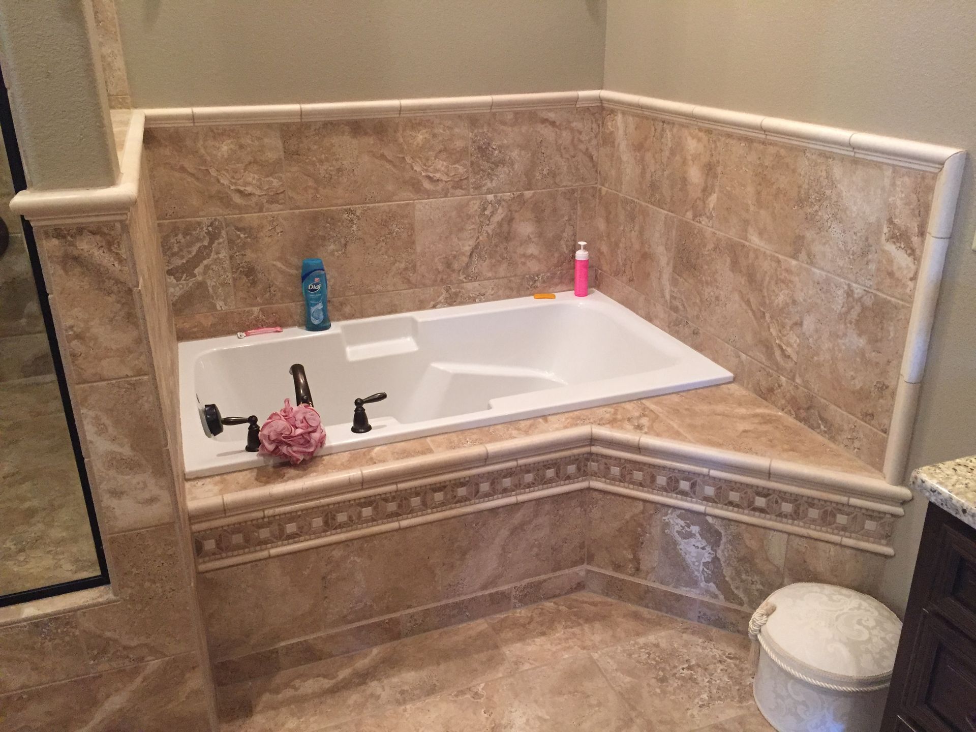 Corner bathtub with beige tile surround, a pink scrub brush, and blue and pink bottles on the ledge.