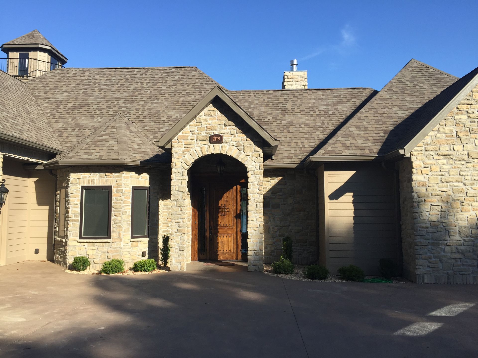 Stone house with arched wooden entry, gray shingle roof, and blue sky overhead