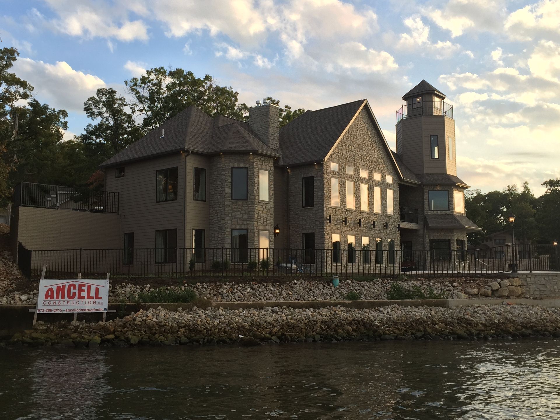 Waterfront stone building with a tower at sunset, beside calm water and a rocky shoreline.