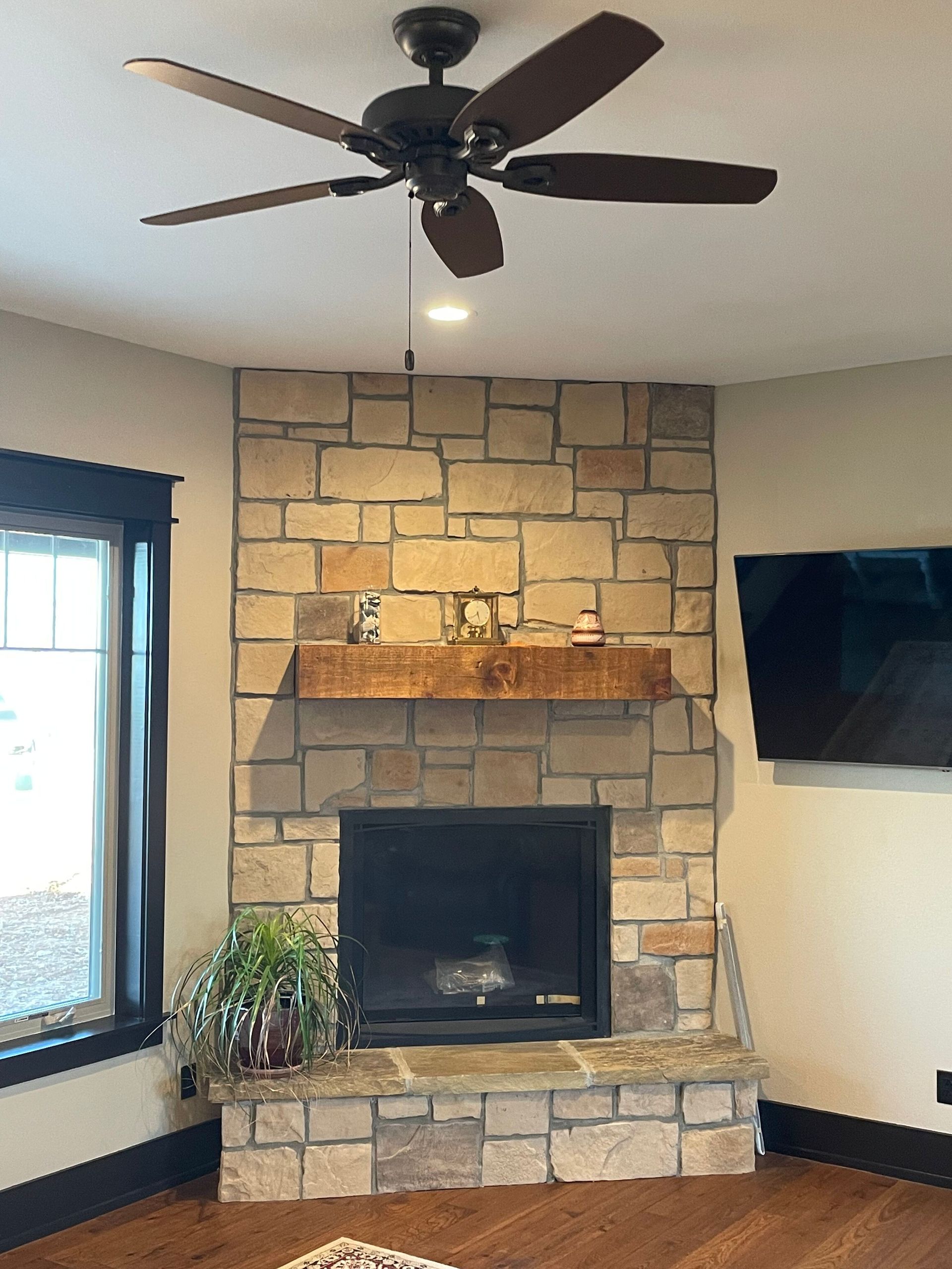 Living room with stone fireplace, mounted TV, ceiling fan, and window