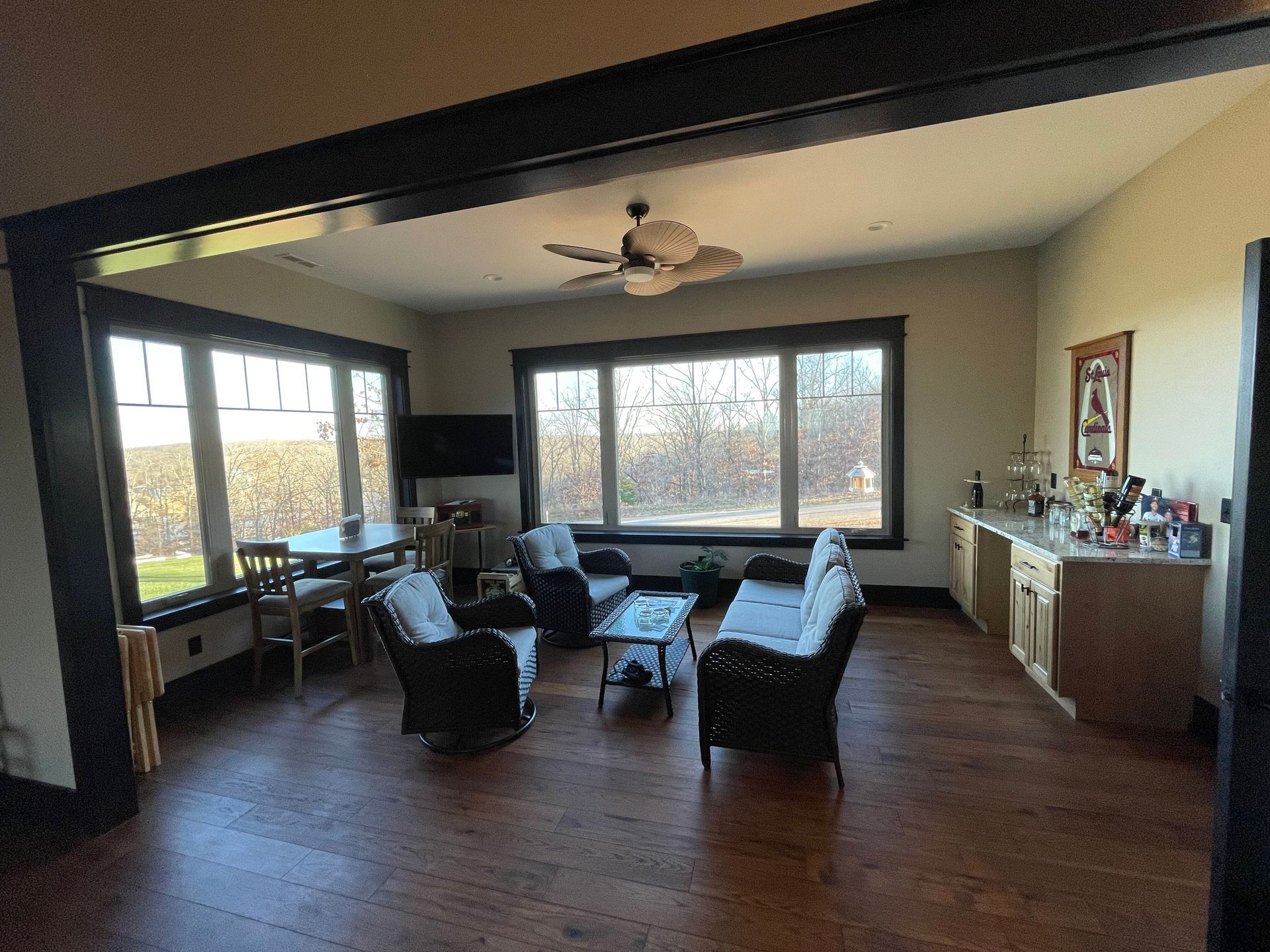 Spacious living room with large windows, dark chairs, a ceiling fan, and a sideboard along the wall.