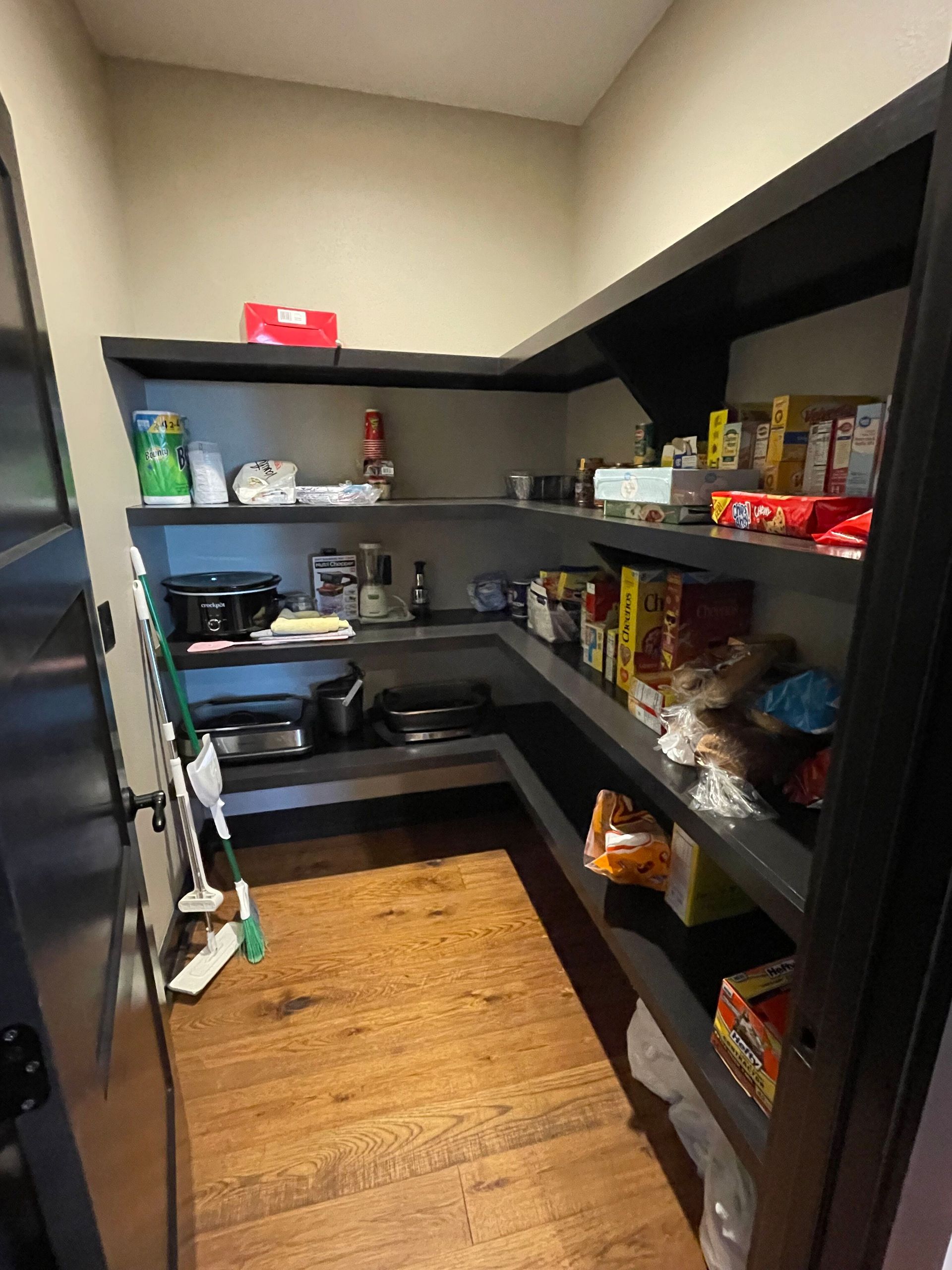 Small pantry with dark shelves, a wood floor, and assorted kitchen items and groceries.