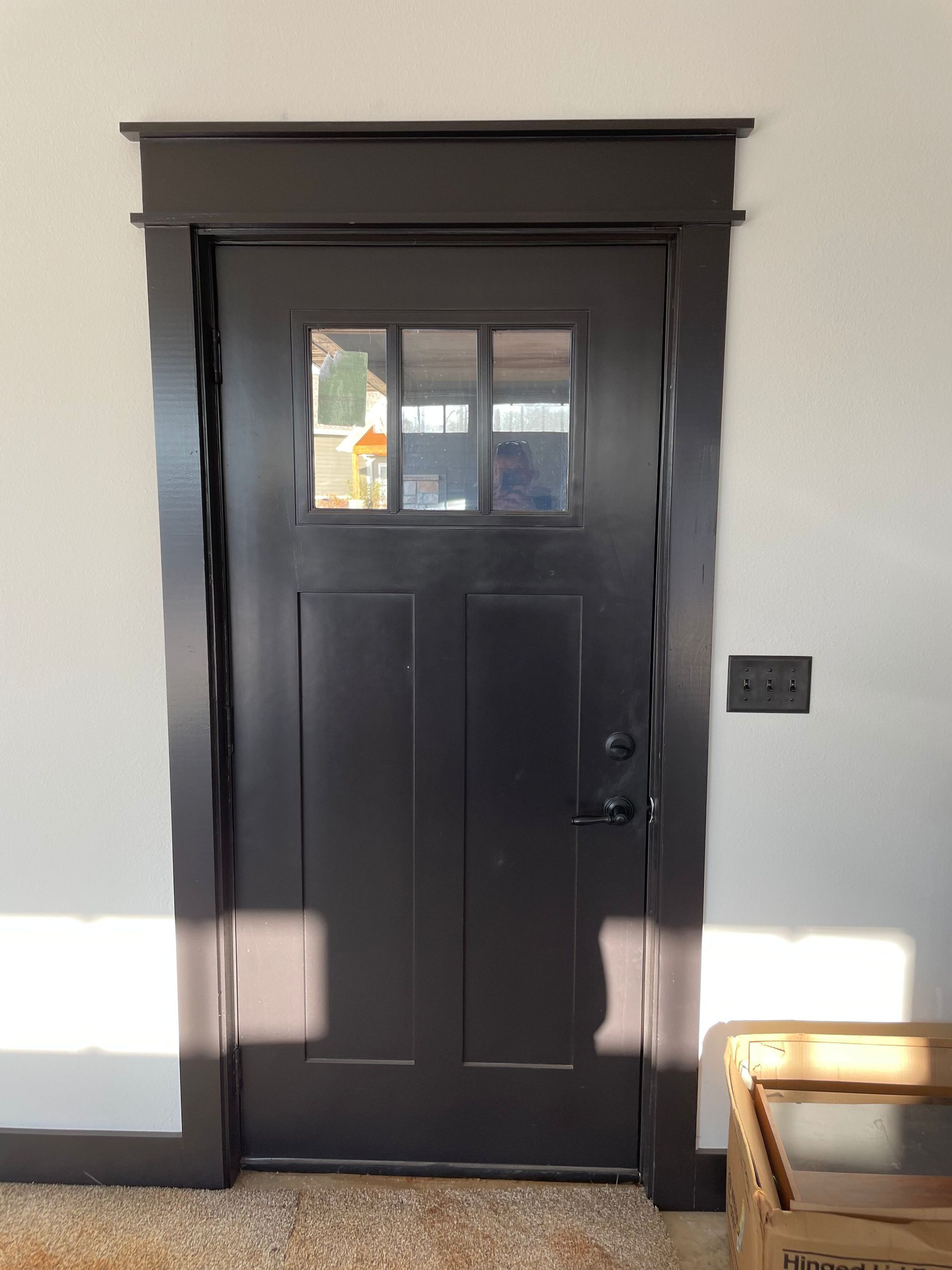 Black front door with small windows in a light gray hallway, with a wall switch and wooden bench nearby