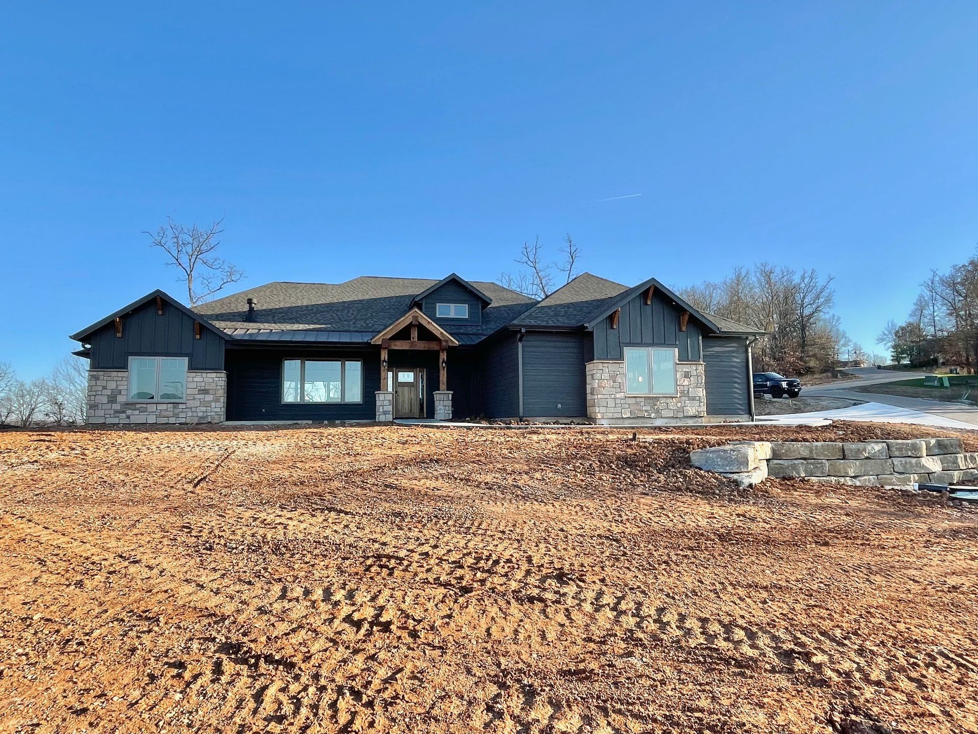 Single-story house with dark roof and blue shutters, set on a bare dirt lot under a clear blue sky