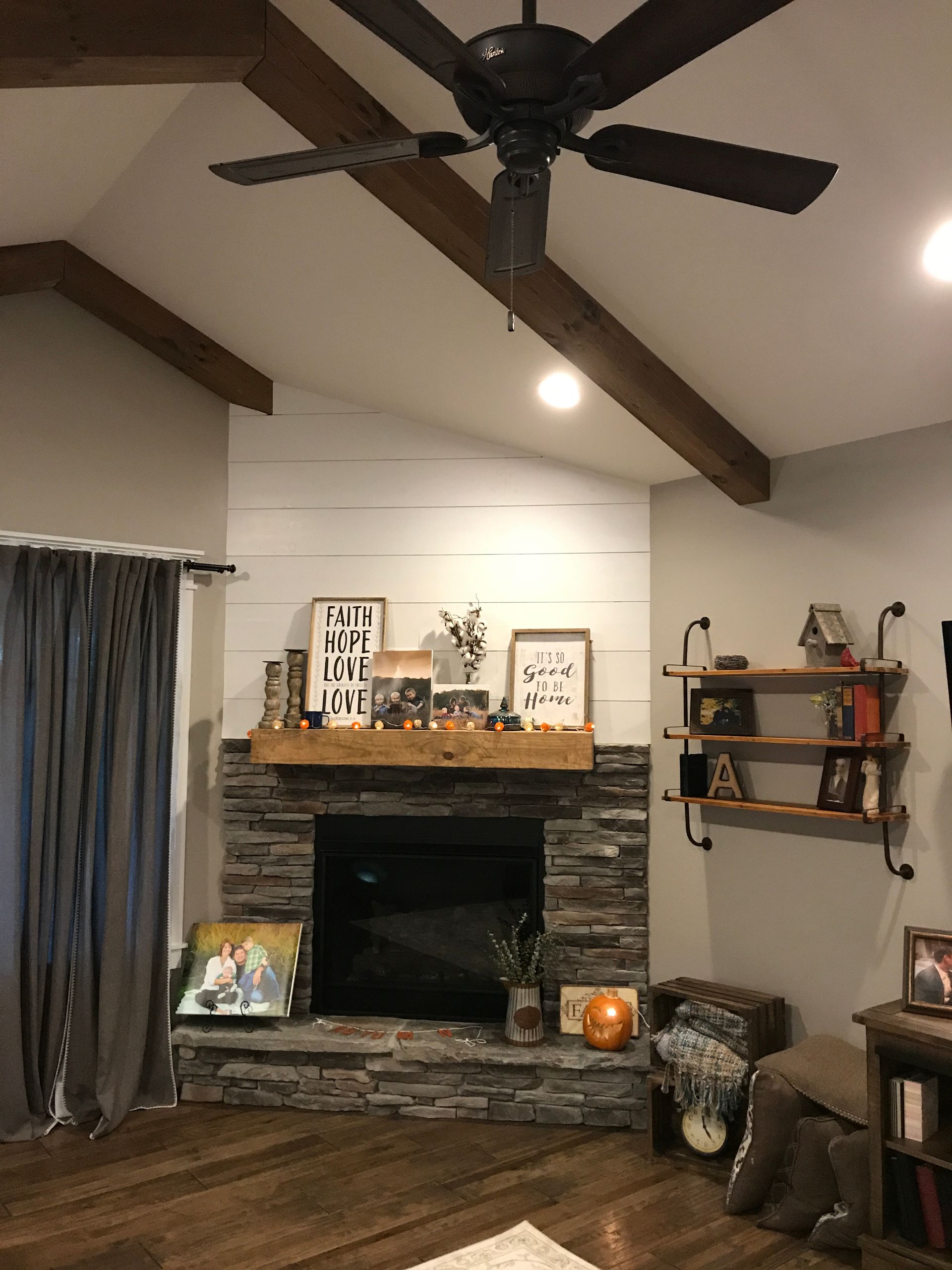 Living room with stone fireplace, ceiling fan, wall shelves, and dark curtains beneath vaulted ceiling